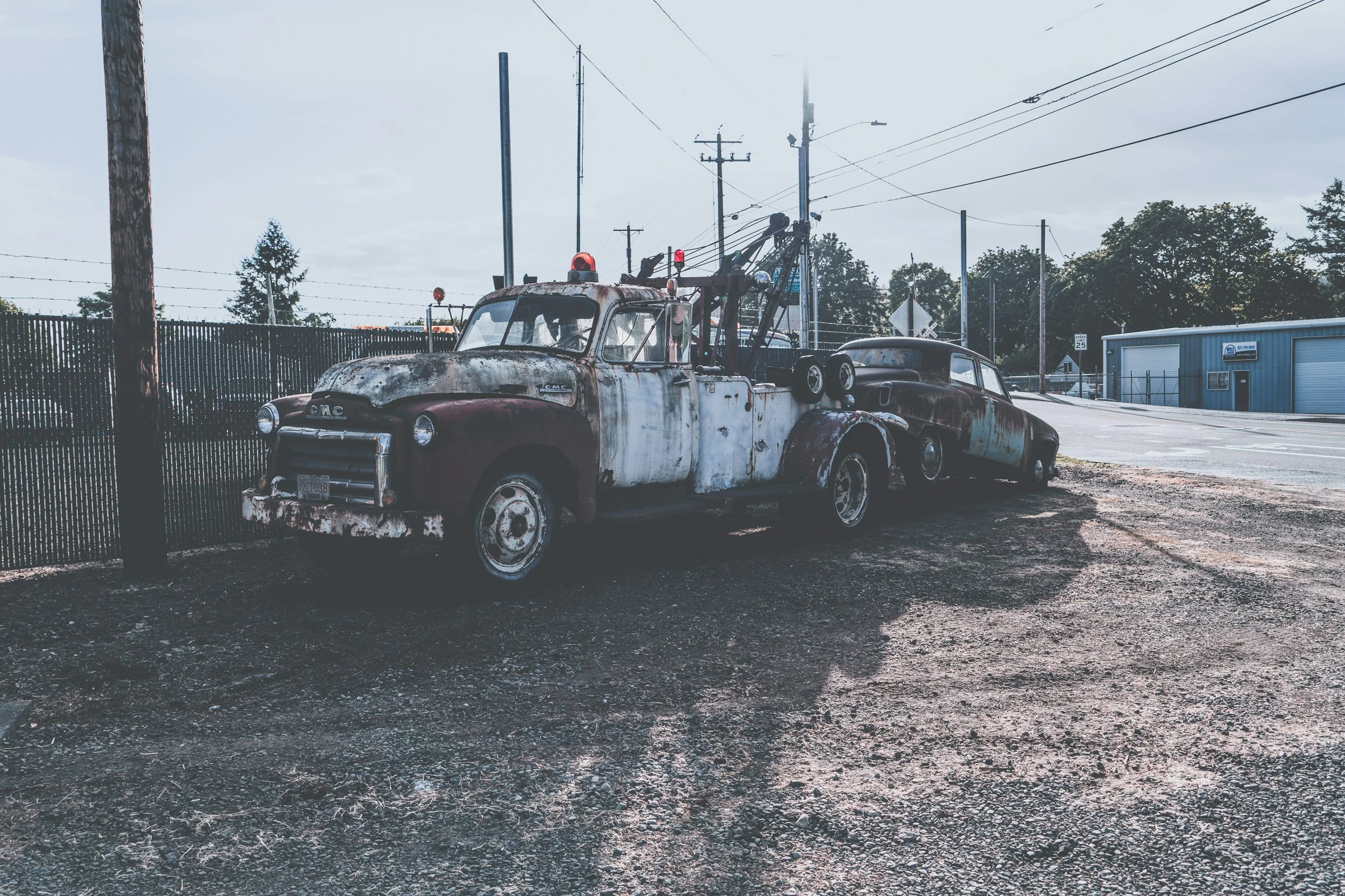 Old rusty tow truck and car parked beside a black metal fence on a dirt lot, with power lines and a blue building in the background.