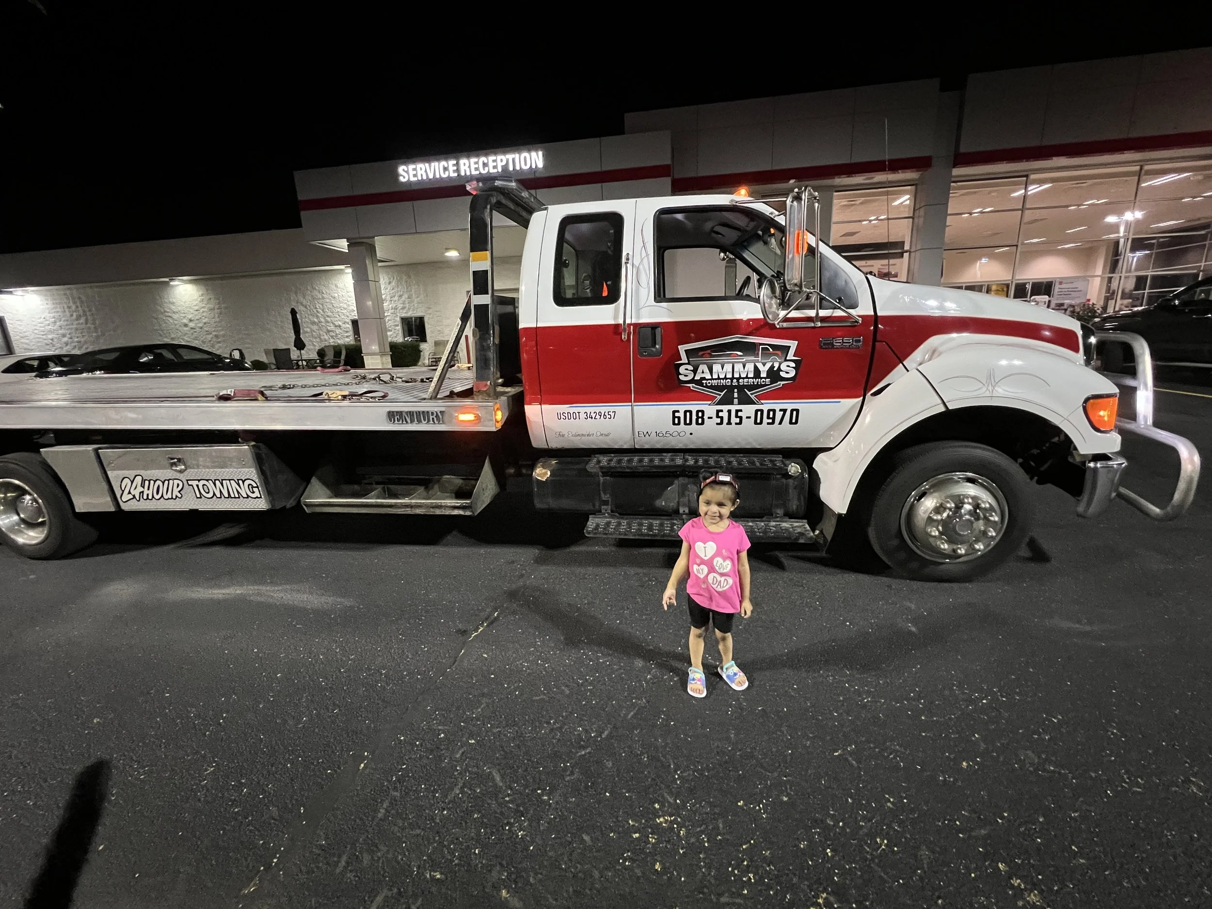A young girl standing in front of a tow truck at night in a parking lot, with a building in the background that has a sign reading 'SERVICE RECEPTION'.