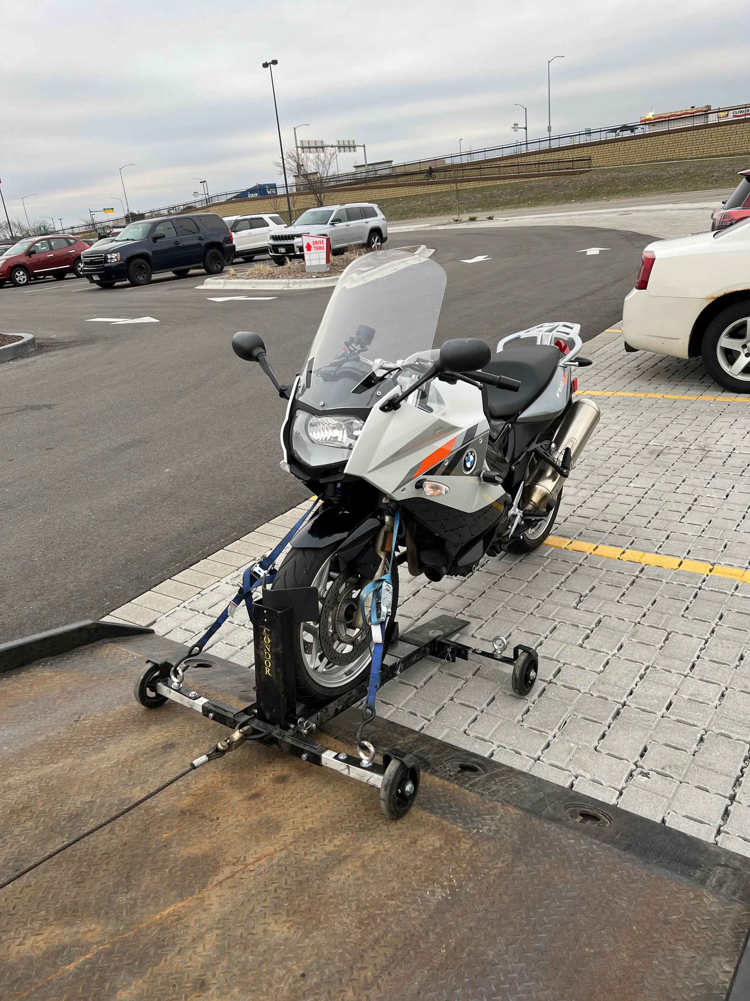A white BMW motorcycle on a motorcycle lift in a parking lot, with other cars and a highway in the background.