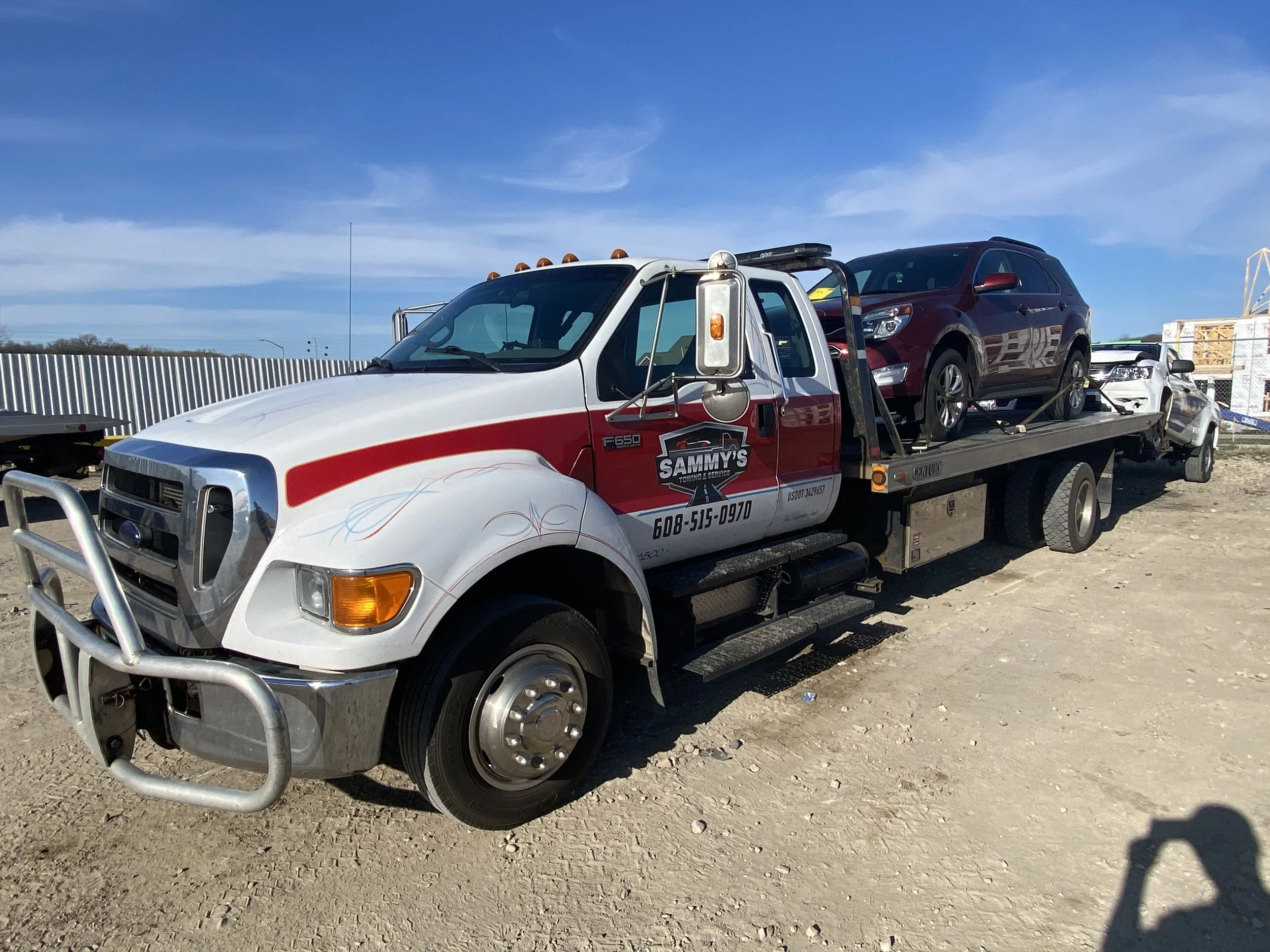 A white and red tow truck with Sammy's towing logo on the door, carrying two cars on its flatbed, parked on dirt under a clear blue sky.