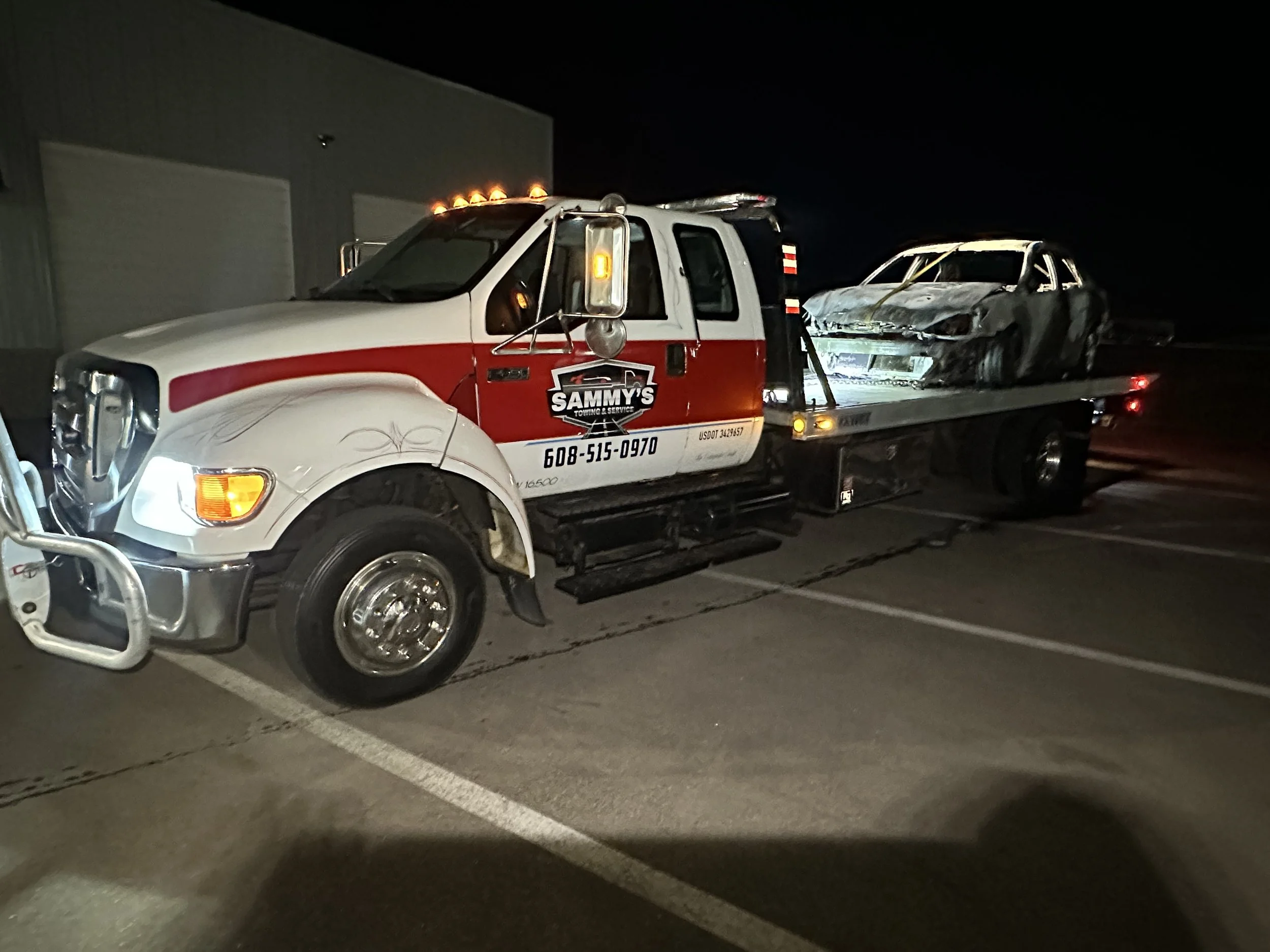 Tow truck with a damaged car on its bed at night in a parking lot.