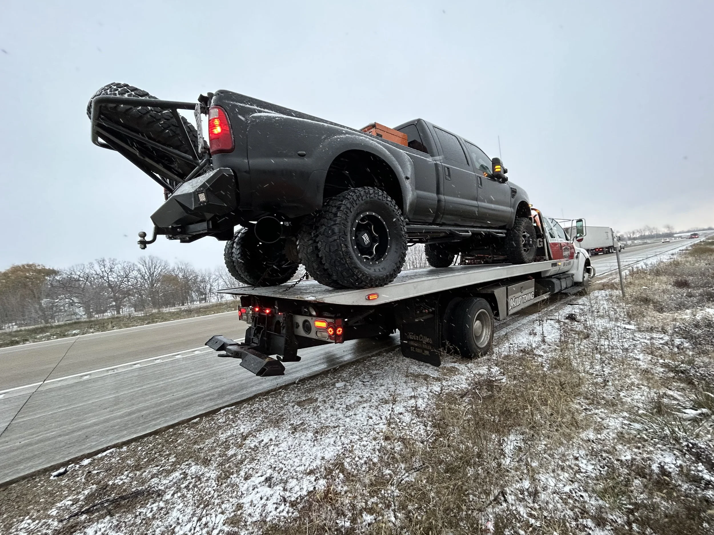 A flatbed tow truck on the side of a highway carrying a black pickup truck during snowy weather.