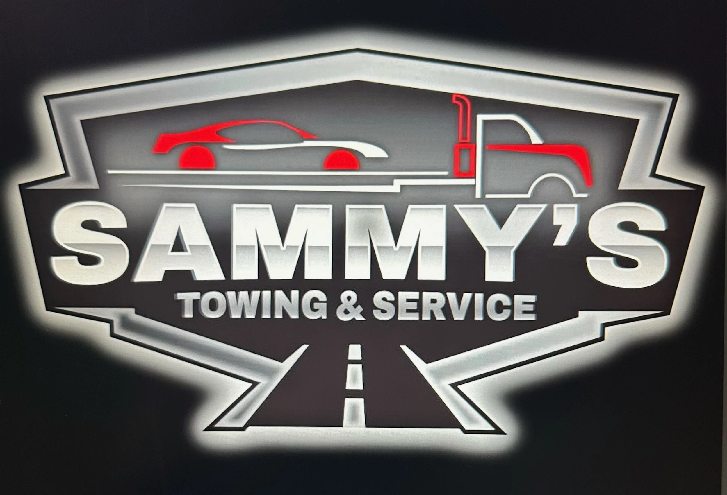 Logo for Sammy's Towing & Service featuring a graphic of a car and a semi-truck, with a road graphic underneath, in black, white, red, and gray colors.
