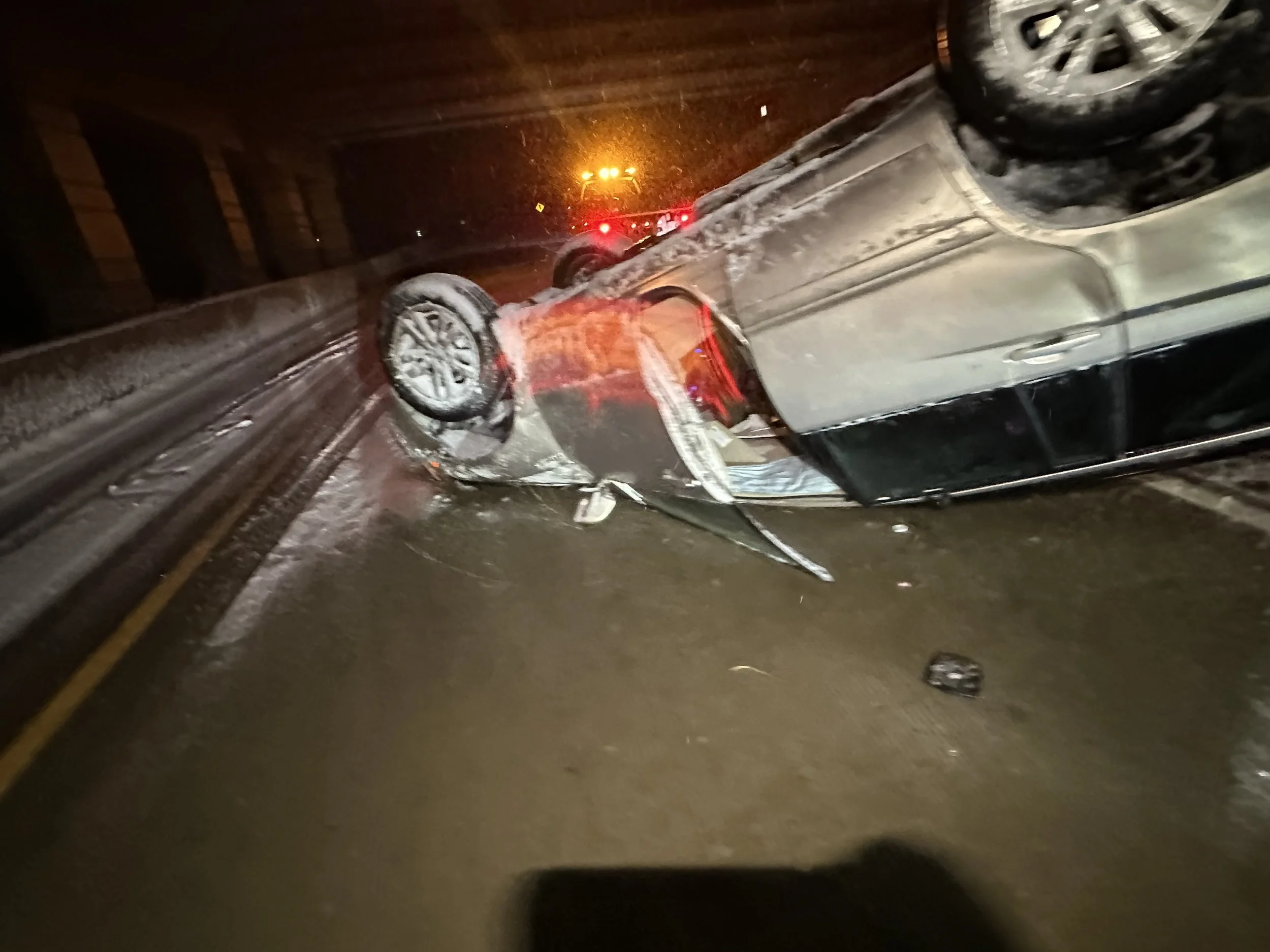 A silver car is flipped upside down on a wet highway, with snow on its roof and tires. Emergency lights are visible in the background.