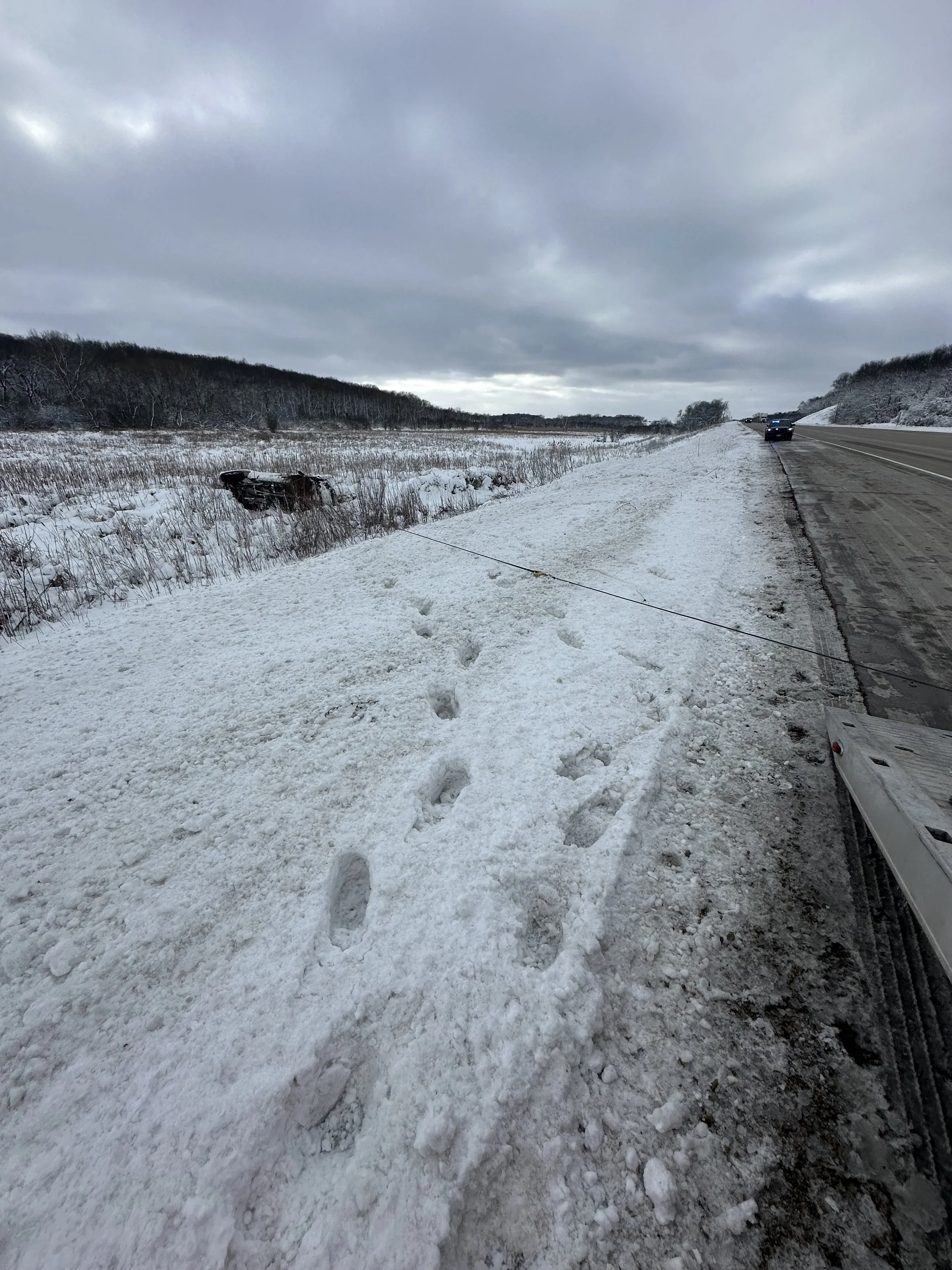 Snow-covered roadside with animal tracks and a tipped-over vehicle in the field, under cloudy sky.