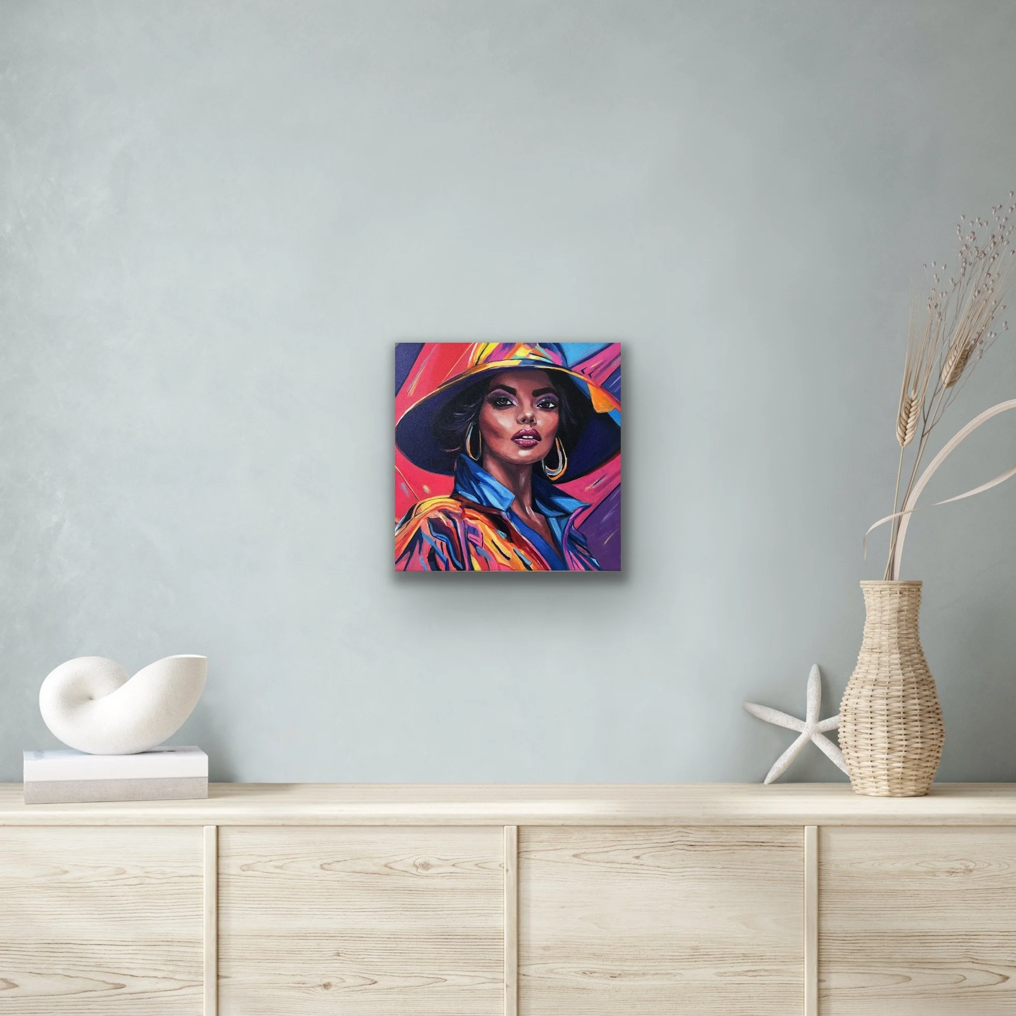 A vibrant portrait of a woman in a wide‑brimmed hat hanging above a light wood cabinet with simple décor.