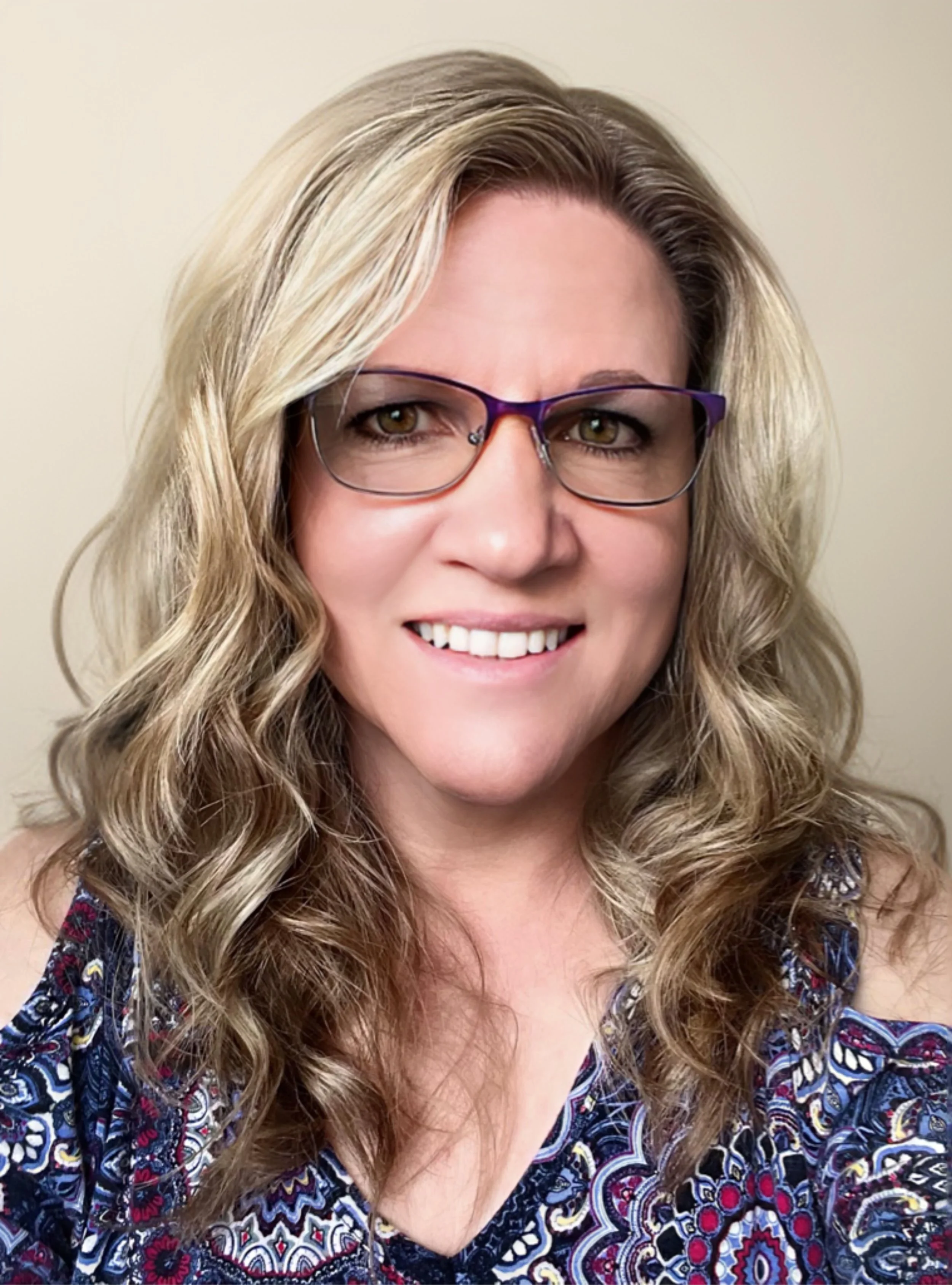 Person with curly blonde hair, wearing eyeglasses and a patterned top, smiling at the camera.