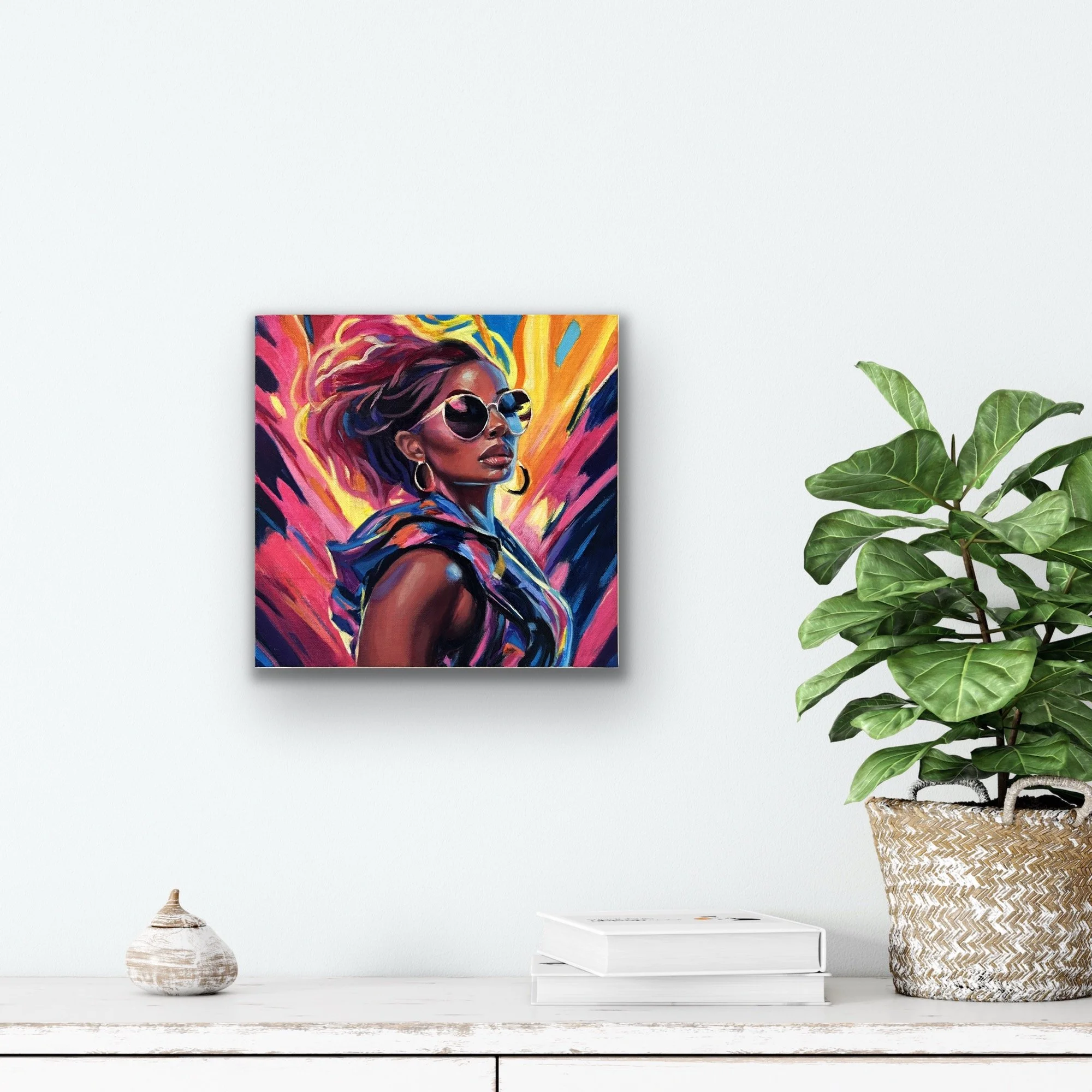 A bold, colorful portrait of a woman in sunglasses and hoop earrings mounted above a white surface with books and a plant.