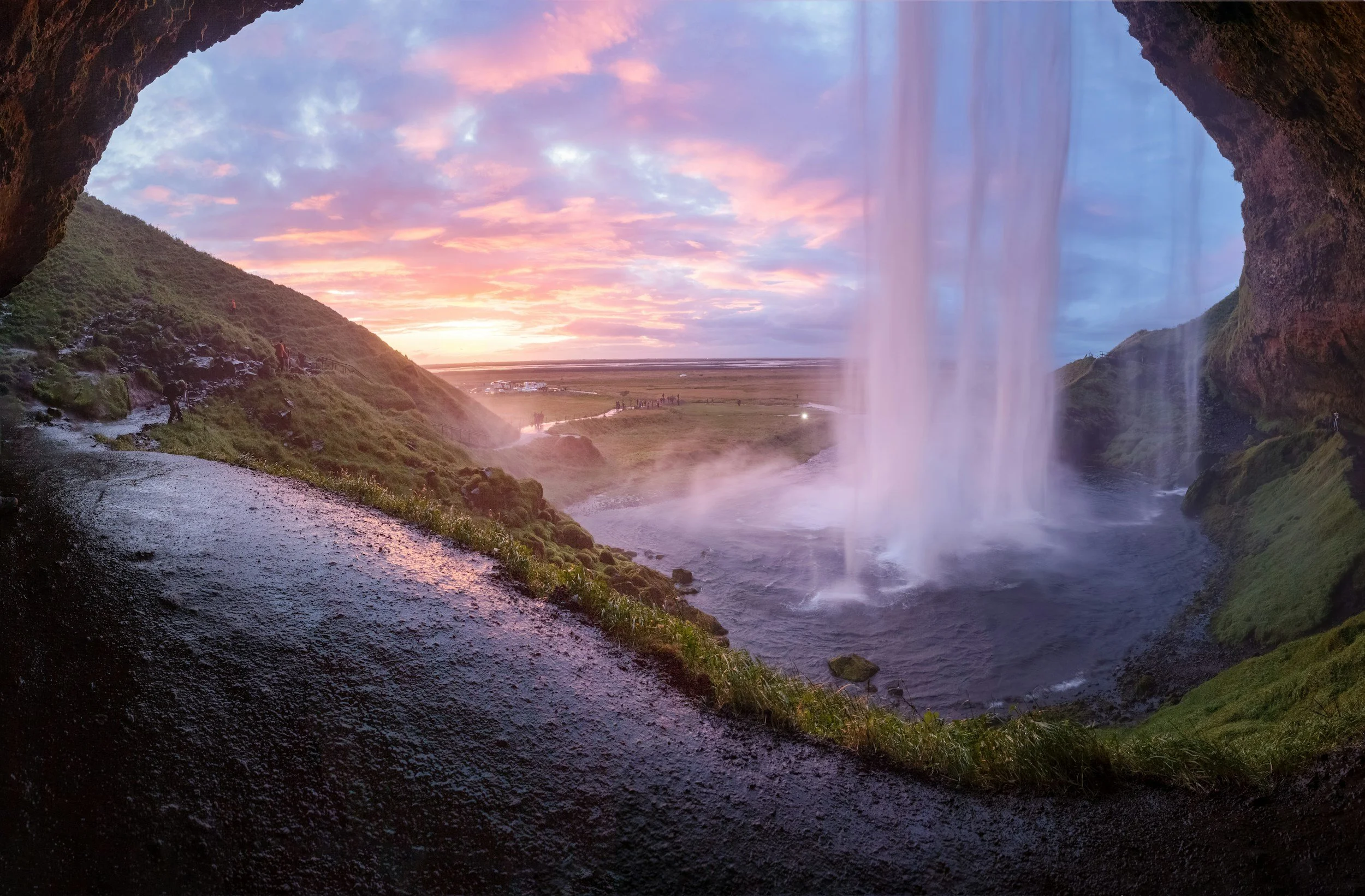 View from behind a waterfall showing a colorful sky at sunset, lush green hills, and a wide open field.