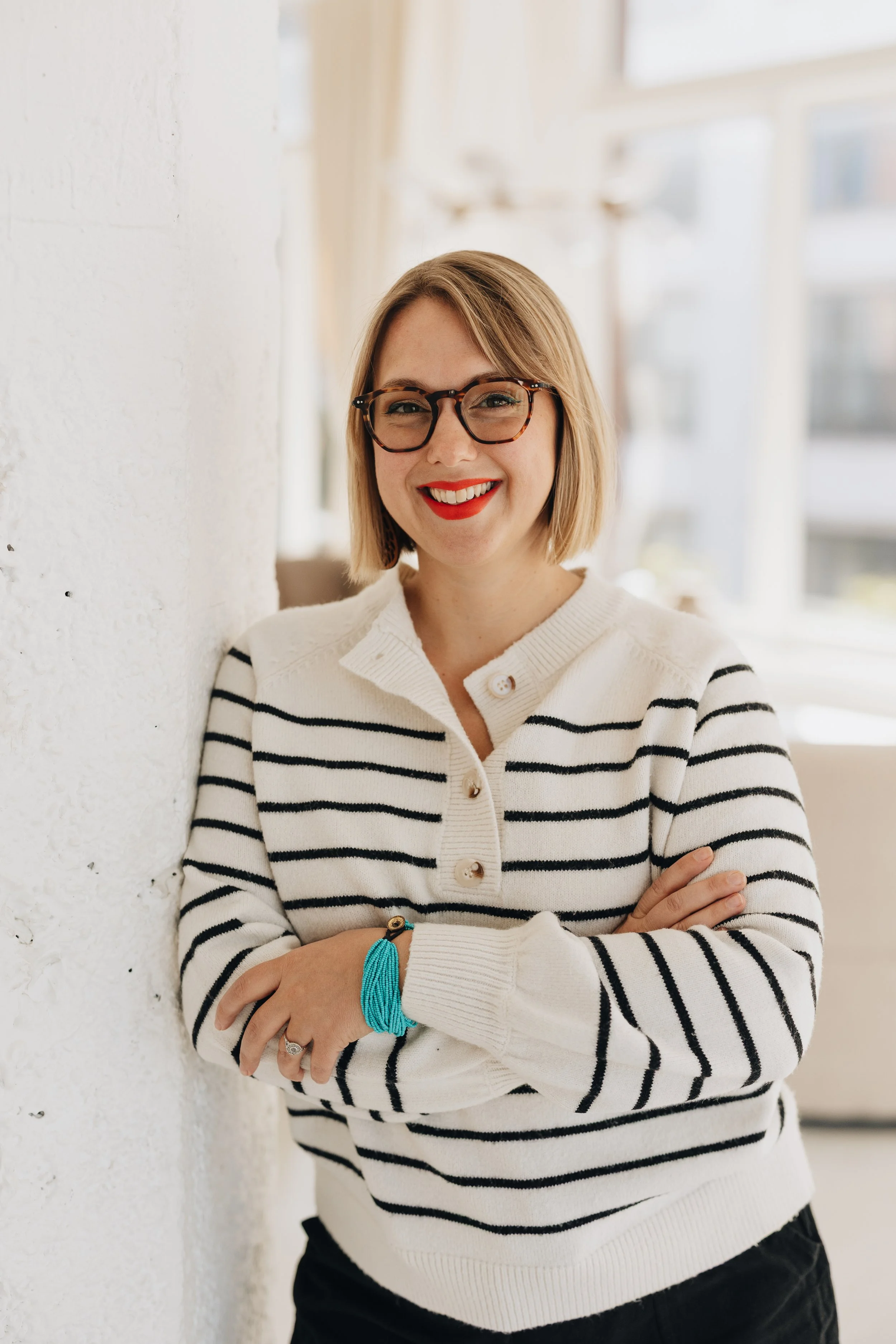 A woman with short blonde hair, wearing glasses, smiling, dressed in a colorful floral top and denim jacket, sitting indoors near a window with potted plants and a brown chair in the background.