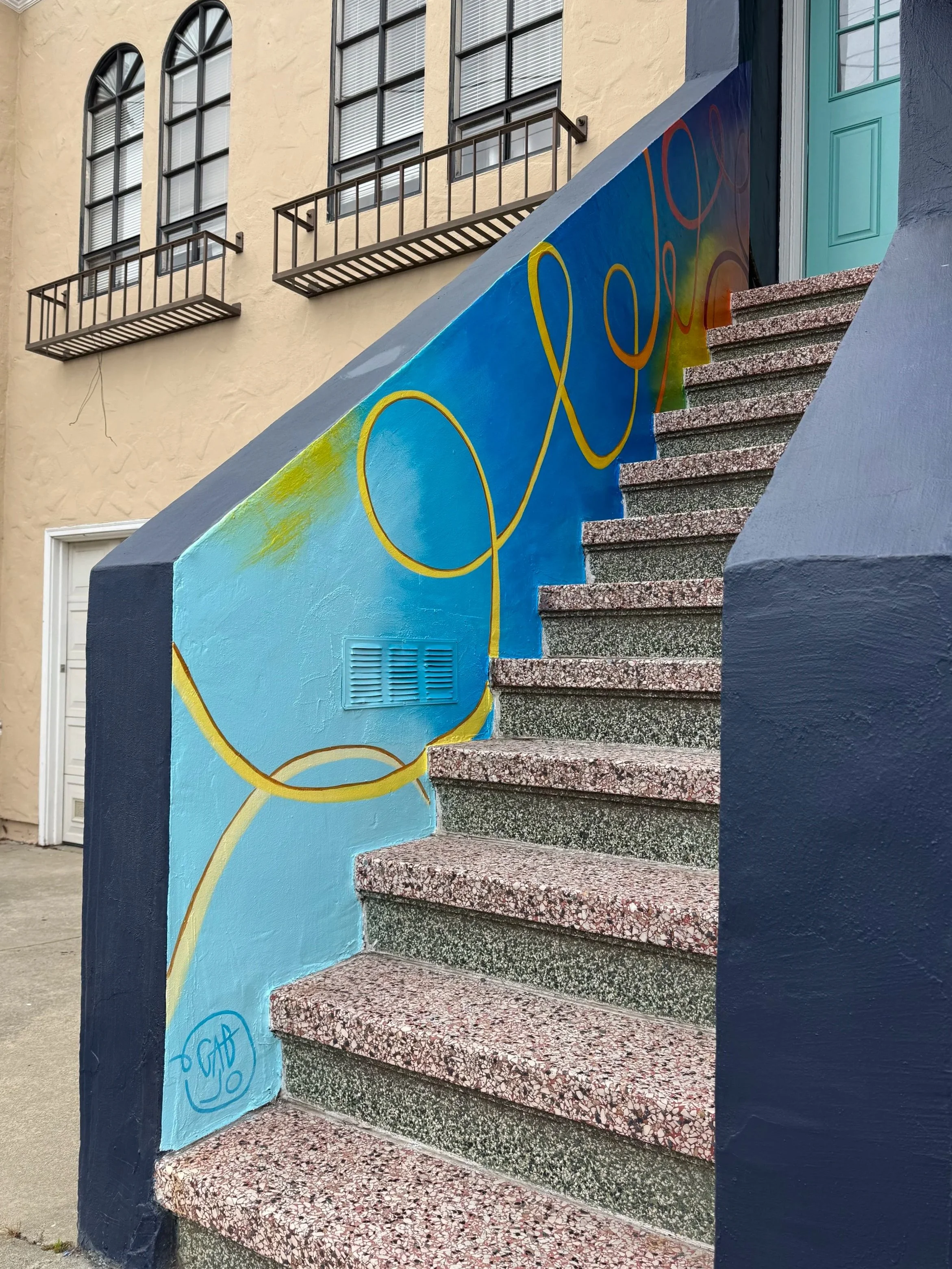 Colorful mural painted on the side of a staircase, with a yellow swirl design on a blue background. The stairs are made of speckled pink and gray terrazzo, leading to a door at the top. The building in the background has beige walls, arched windows with black metal railings, and a teal door.