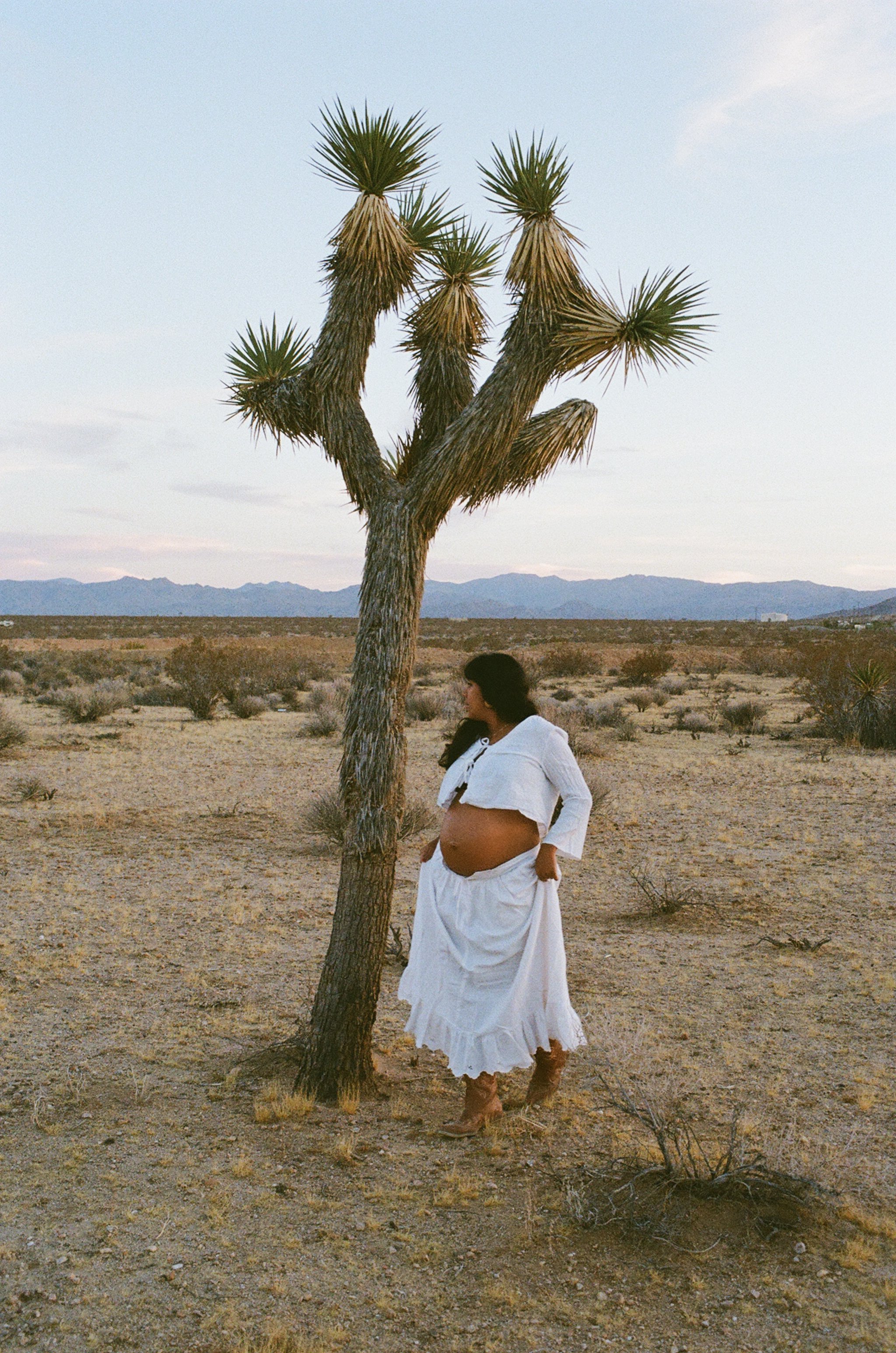 Pregnant GabJo in a white dress and shawl standing in a desert landscape with a Joshua tree and mountains in the background.