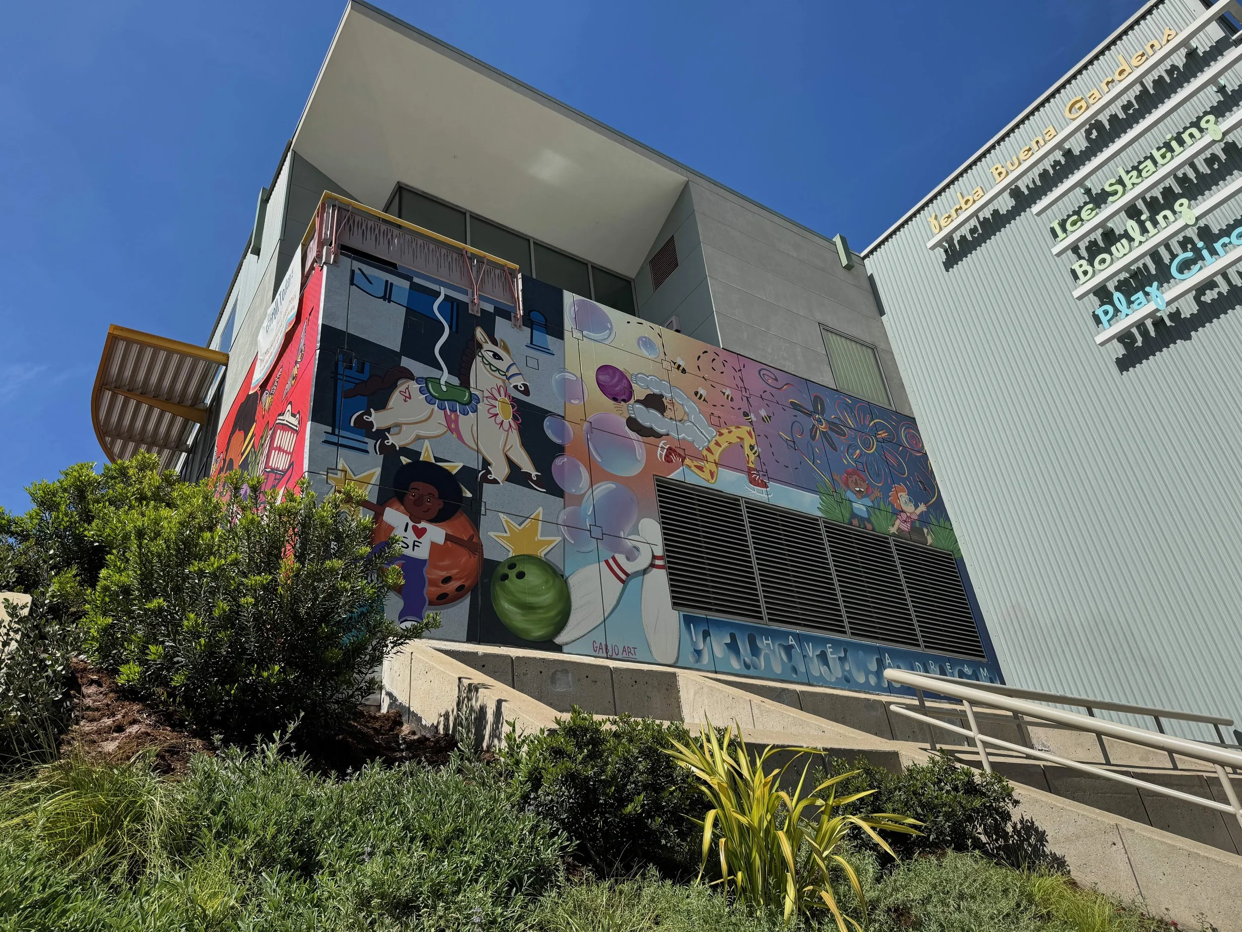 Colorful mural on the side of a building featuring various cartoon characters, animals, and whimsical elements, with a landscape of bushes and plants in the foreground.