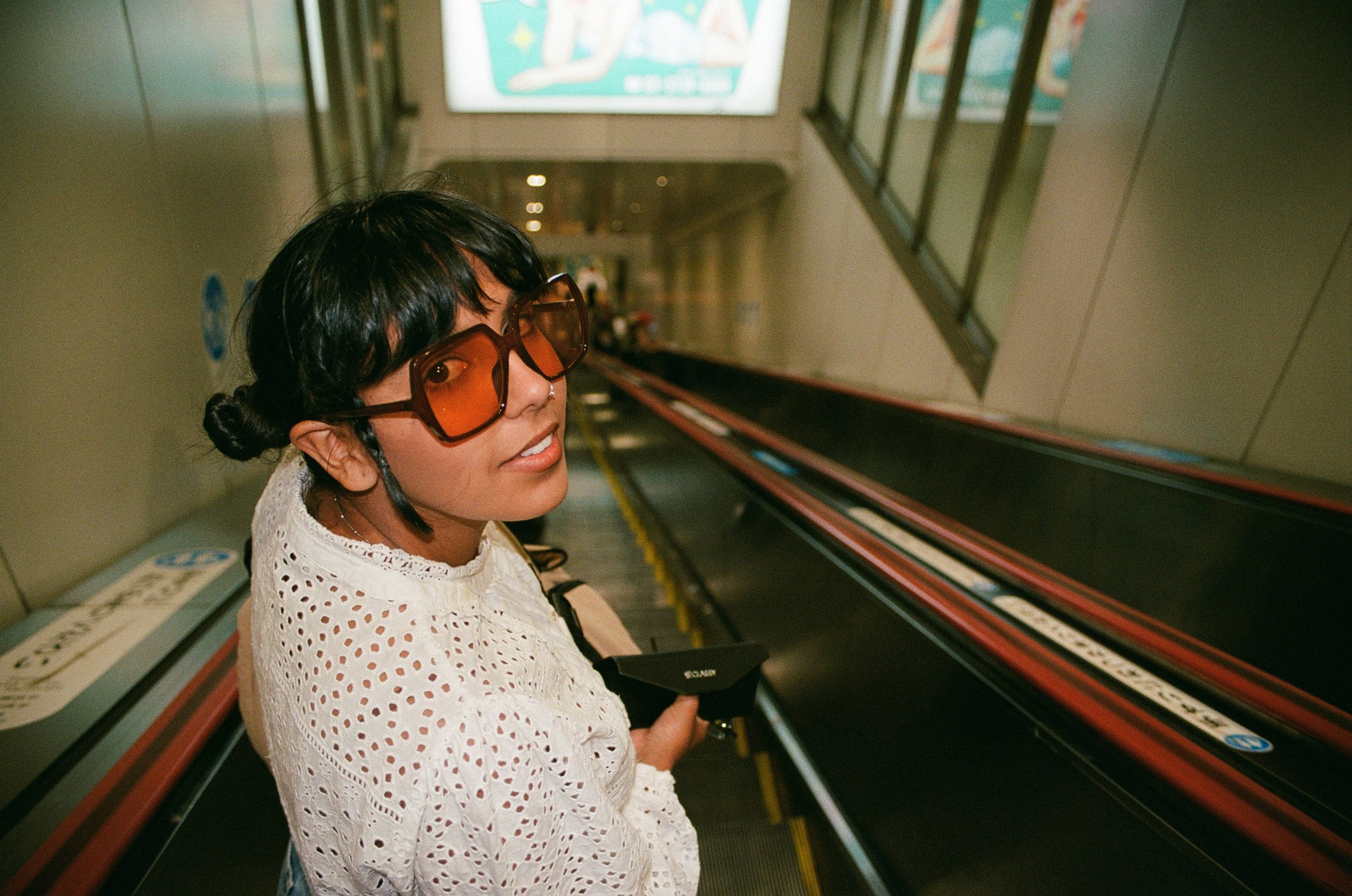 GabJo with short dark hair wearing large orange sunglasses and a white eyelet lace top riding an escalator in an indoor setting.