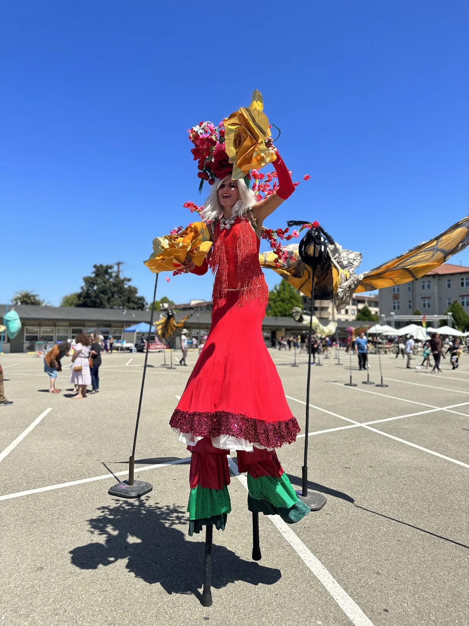 An artist dressed in a vibrant, elaborate costume rides on stilts at an outdoor event. The costume features a long red dress with decorative trim, large shoulder accents made of yellow fabric, and a headpiece with bright pink flowers. There are peopl