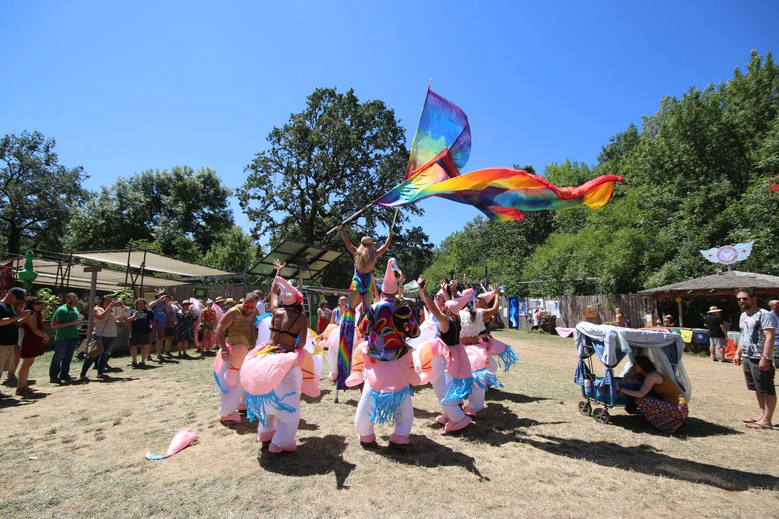 People at an outdoor festival dancing in costumes, with one person waving a rainbow flag, under a clear blue sky, surrounded by trees.