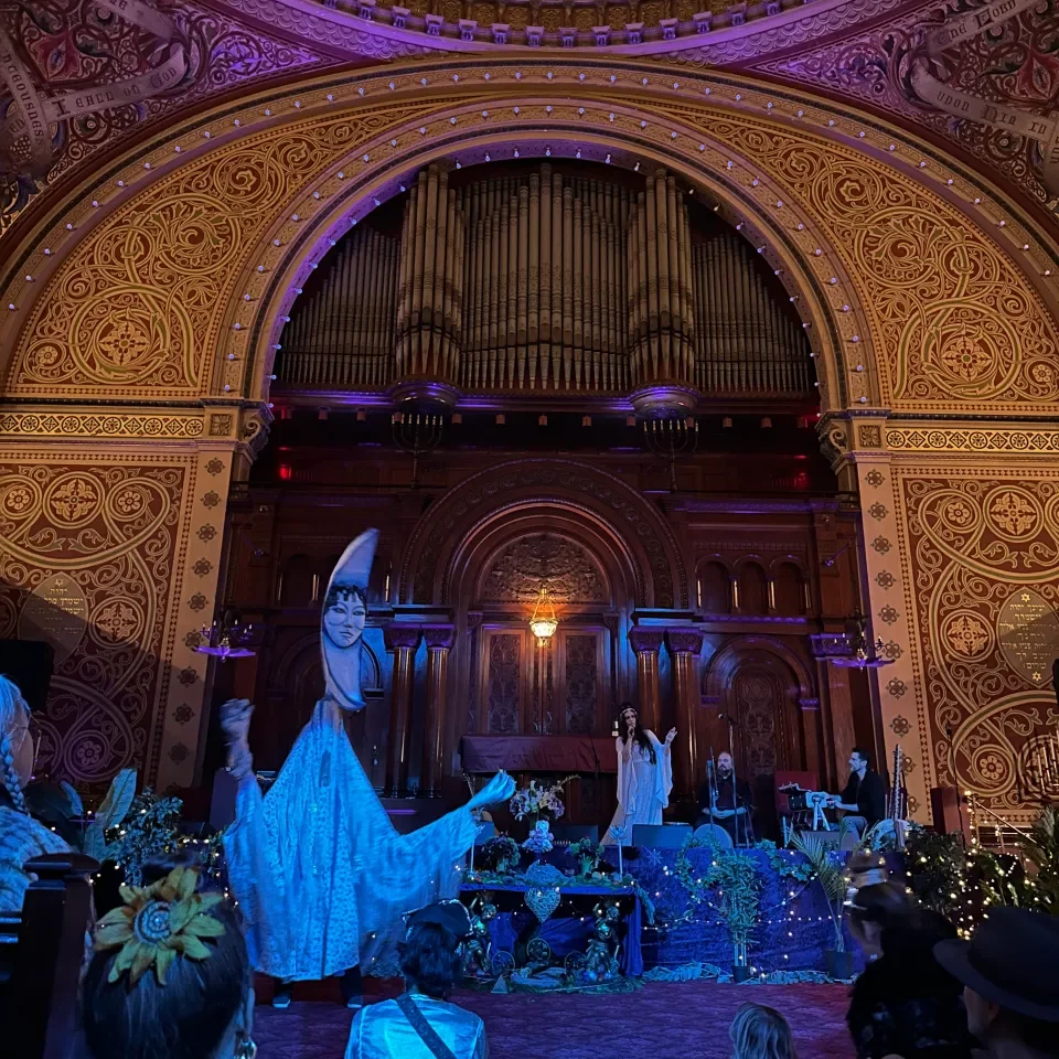 An indoor stage set up for a performance inside a decorated church or auditorium with ornate gold and red interior, featuring a pipe organ at the back, a person in a white dress performing, and audience members watching.