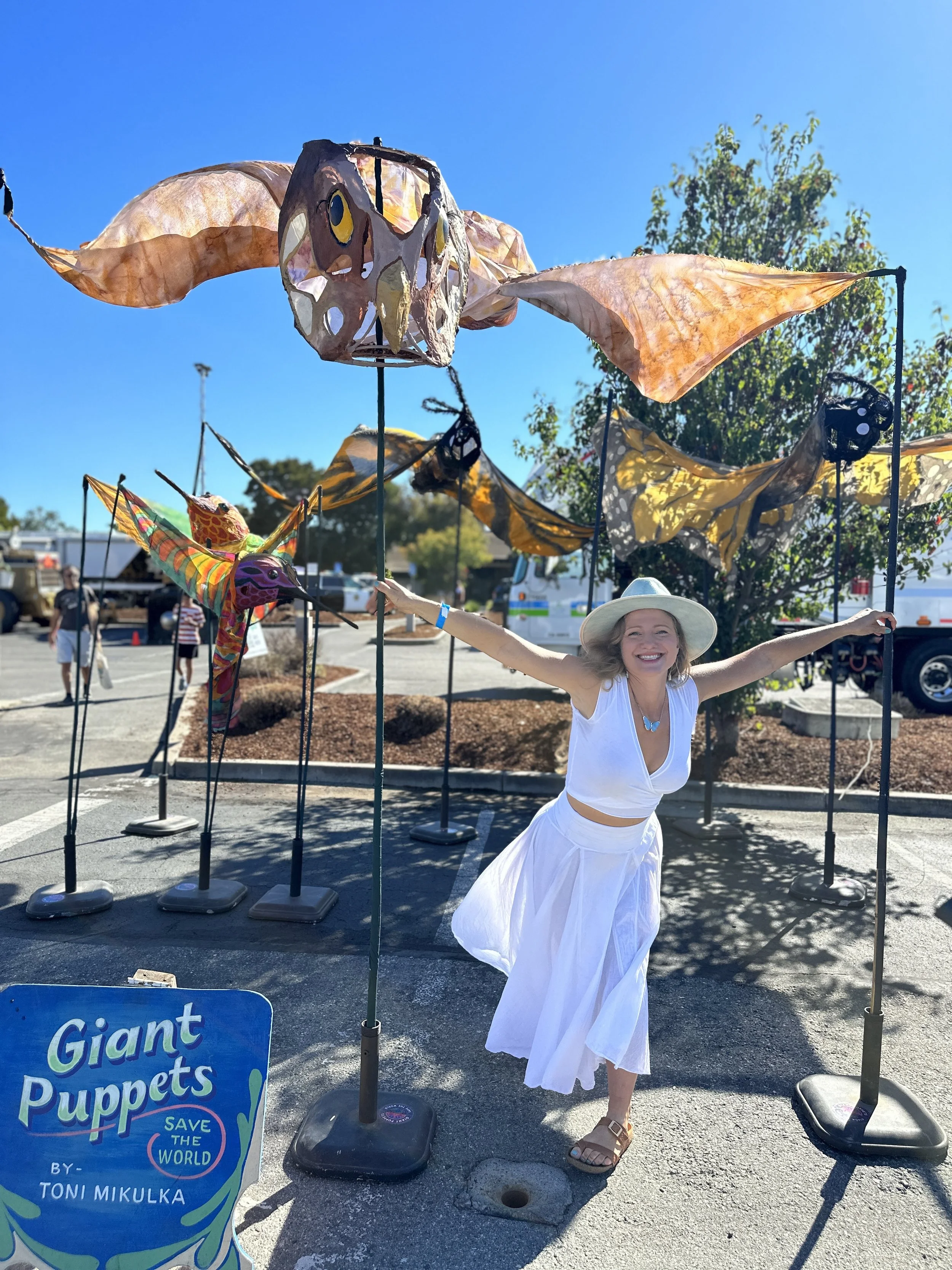 A woman in a white dress and wide-brimmed hat stands in a parking lot, smiling and holding up giant moth and butterfly puppets at an outdoor art event, with other puppets and people in the background.