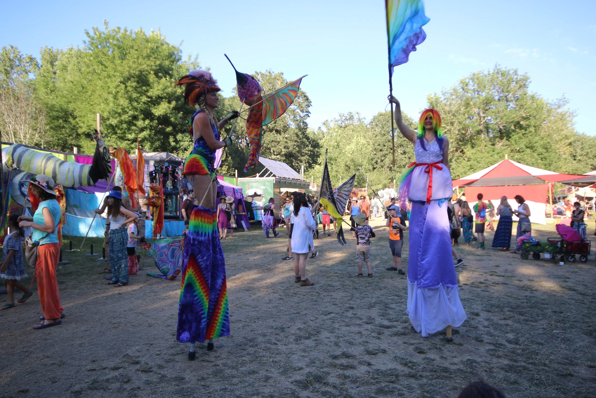At a lively outdoor festival, two performers on stilts wearing colorful costumes and rainbow wigs entertain a crowd. The performers are holding rainbow flags and are surrounded by festival attendees, some of whom are also wearing colorful clothing an