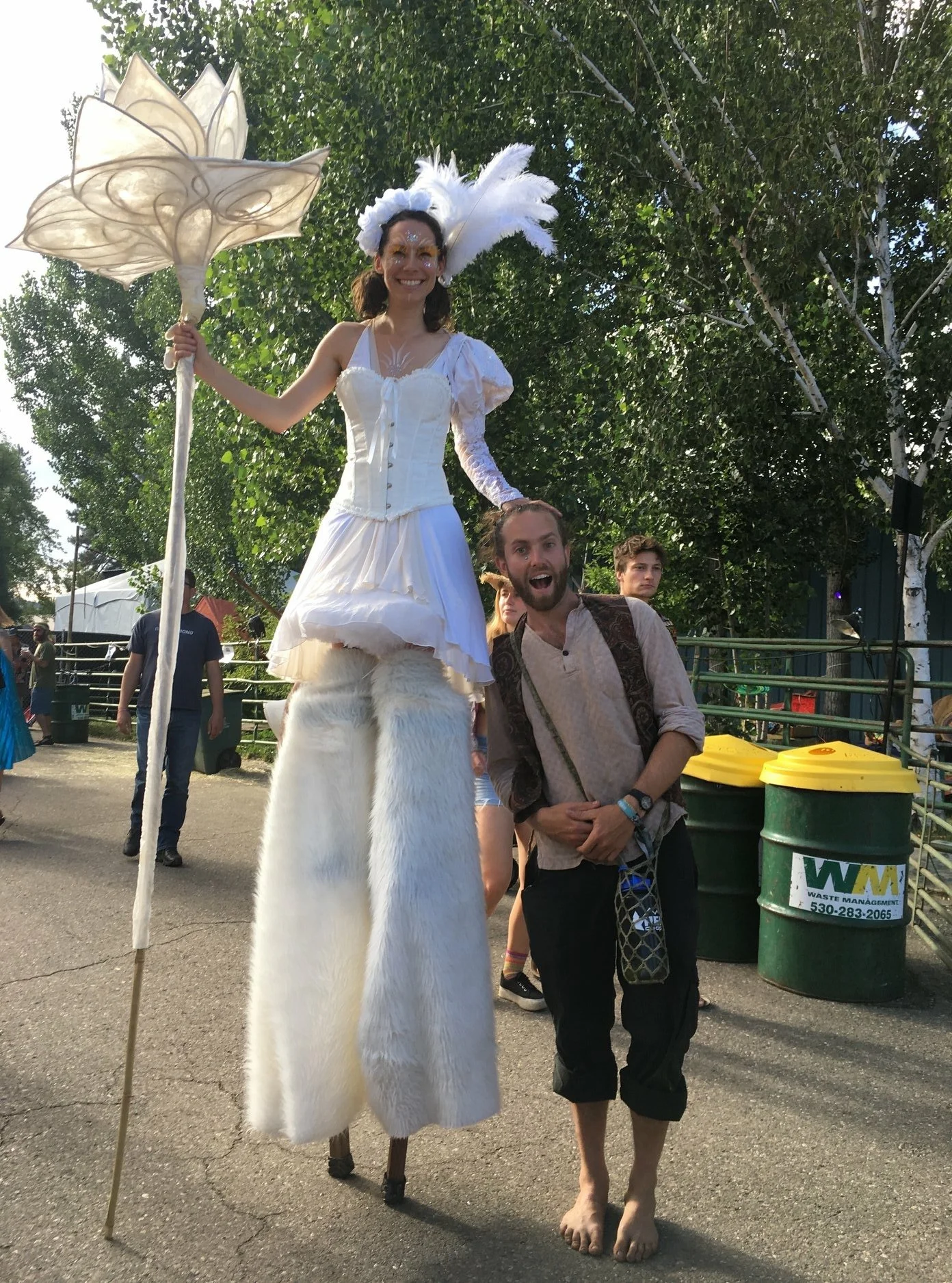 A woman dressed as a fairy godmother standing on stilts, wearing a white dress and feathered accessories, holding a staff, and smiling at a man standing next to her during an outdoor event.