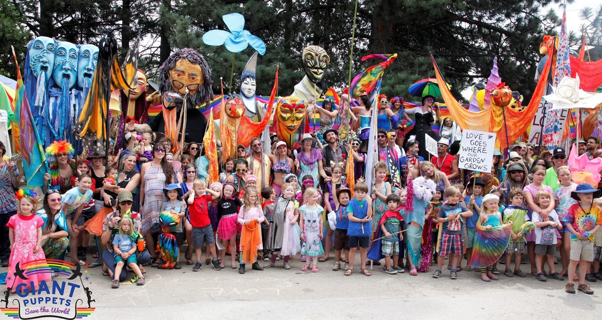A large group of people, including children and adults, gathered outdoors during a festival or parade, holding various colorful flags and signs. Many participants are dressed in vibrant, rainbow-colored clothing and costumes, with some wearing masks 
