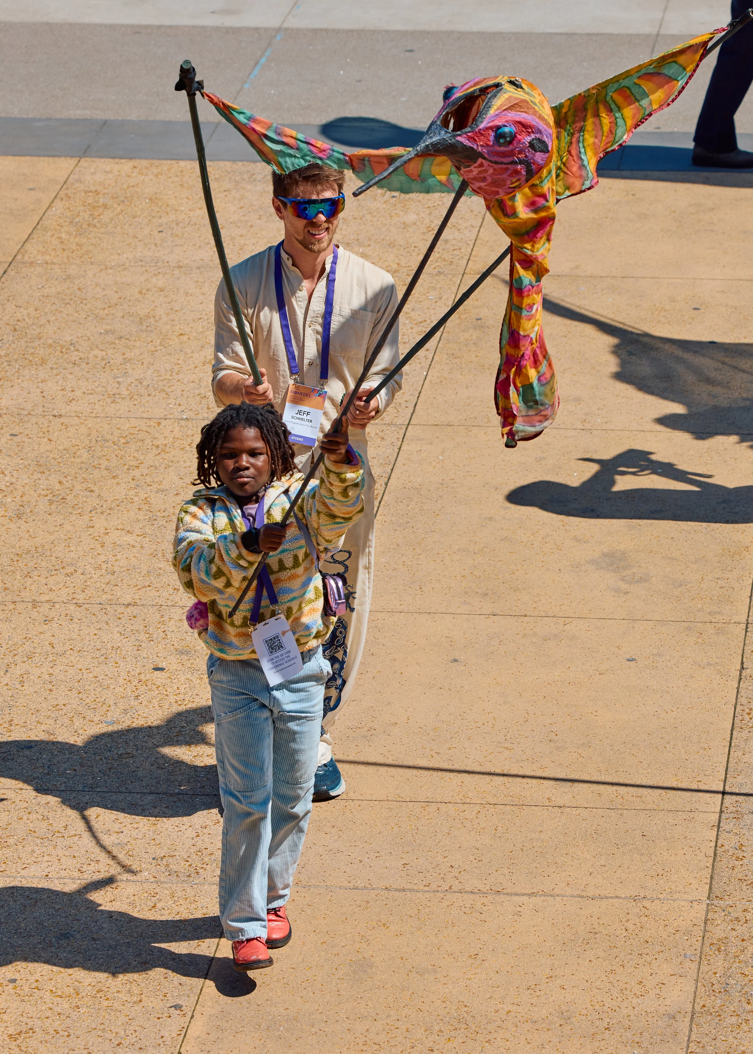 A man and a young girl holding a large, colorful, bird-shaped kite with dragonfly-like wings, flying it outdoors on a sunny day.