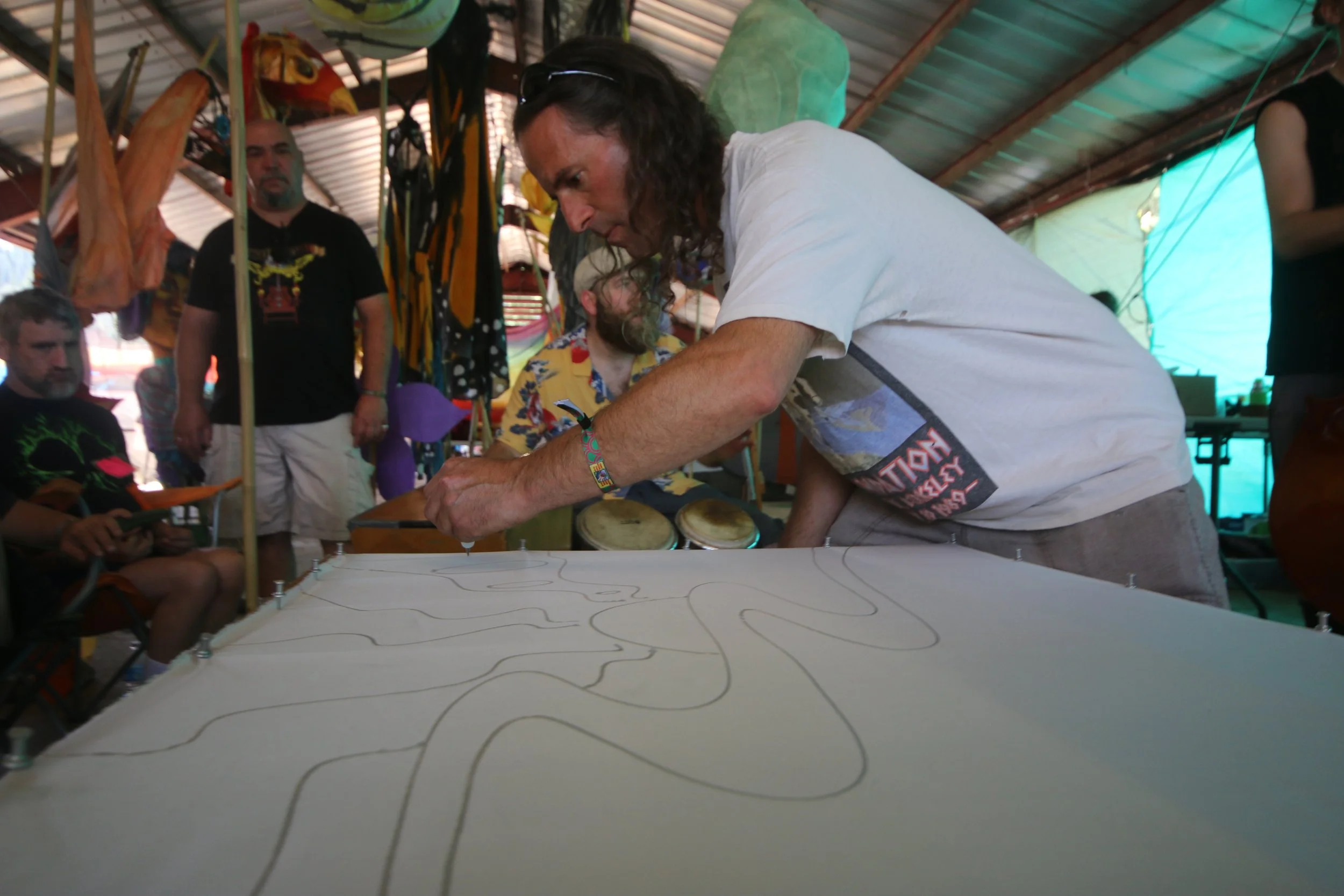 A man with long dark hair and a white t-shirt leaning over a large white surface, drawing on it with a thin tool, while a group of people watches inside a colorful tent or shelter.