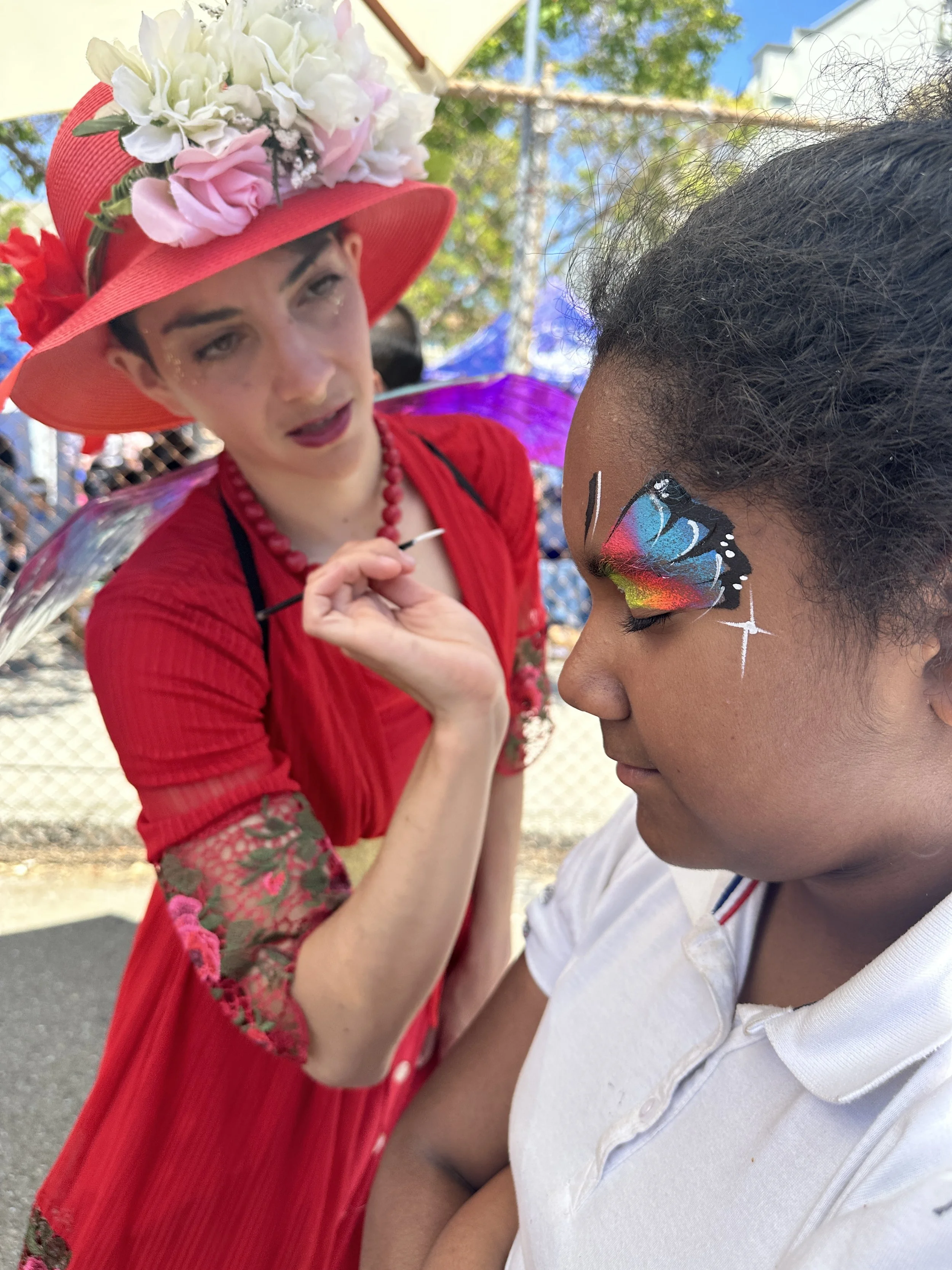 A woman dressed in red with a floral hat is painting a butterfly design on a young girl’s face at an outdoor event.
