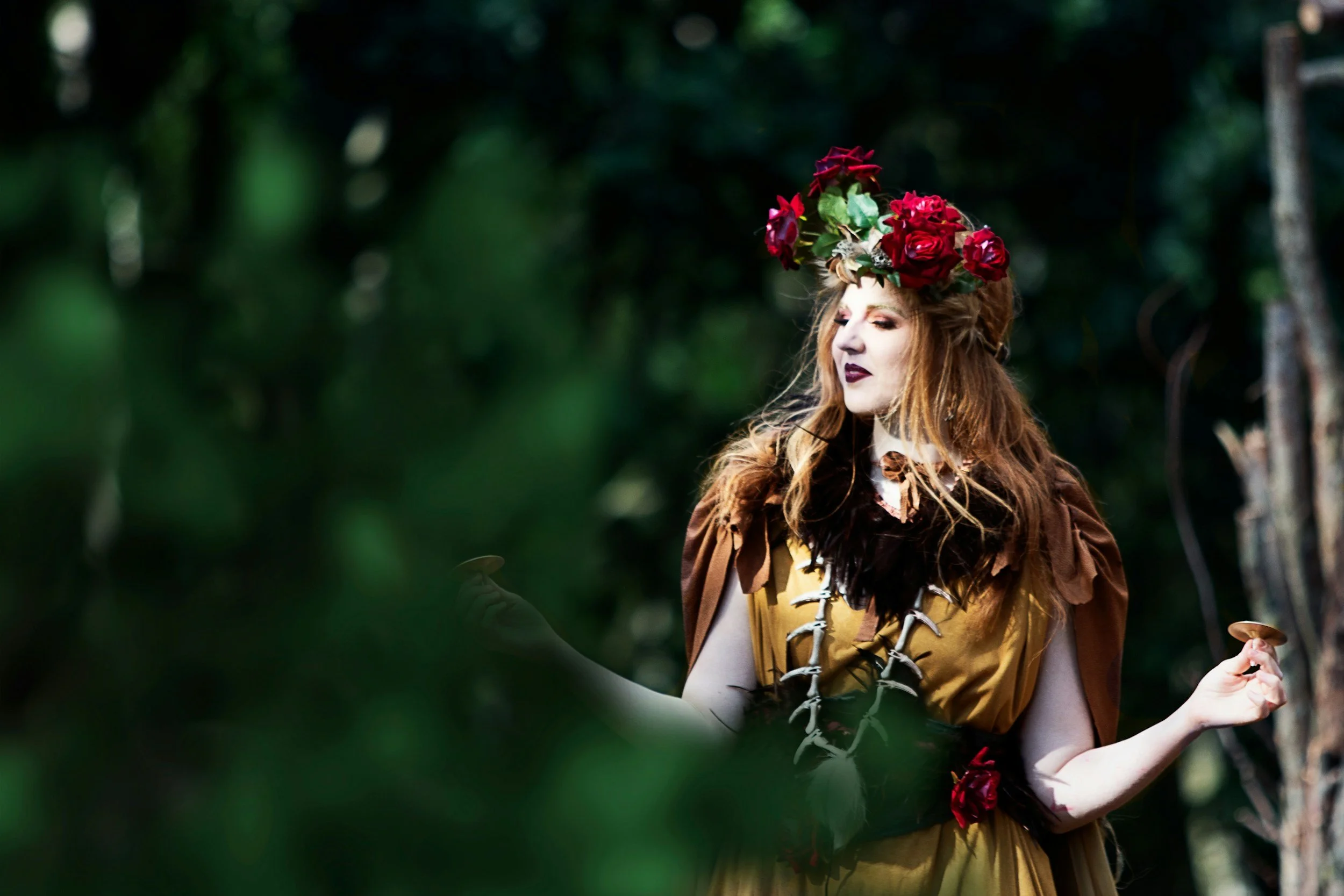 A woman dressed as a fairy or mythical character with a crown of red roses, wearing a brown and yellow dress with feather and bone details, standing in a forest with a mystical or fantasy appearance.
