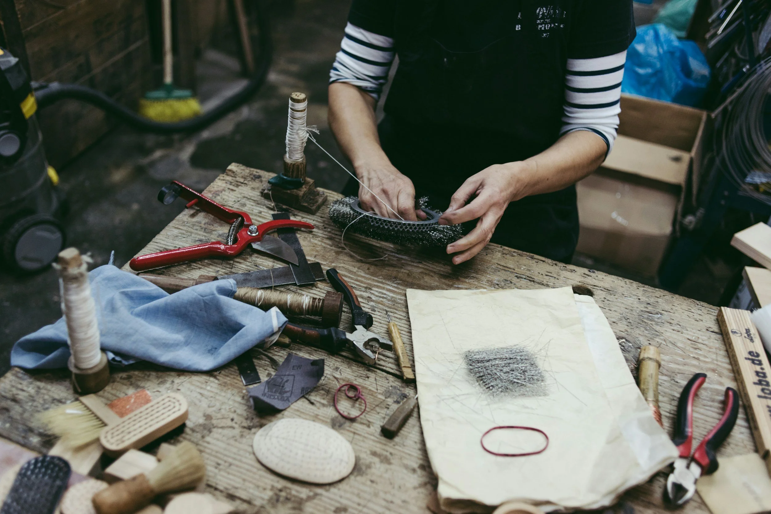 Person working at a cluttered wooden workbench with tools, threads, and various materials, crafting or repairing an object.