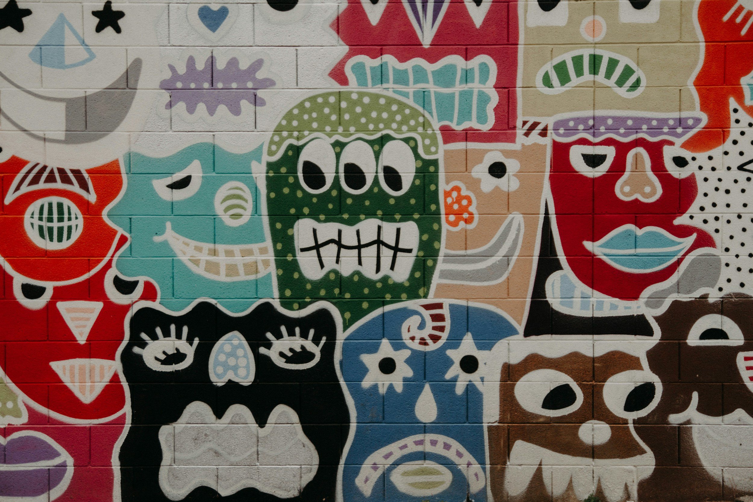 Colorful mural with stylized cartoon faces, including a green skull with three eye holes, a blue face with star-shaped eyes, a red face with lips and eyelashes, a black face with white eyes and mouth, and various abstract shapes in vibrant colors.