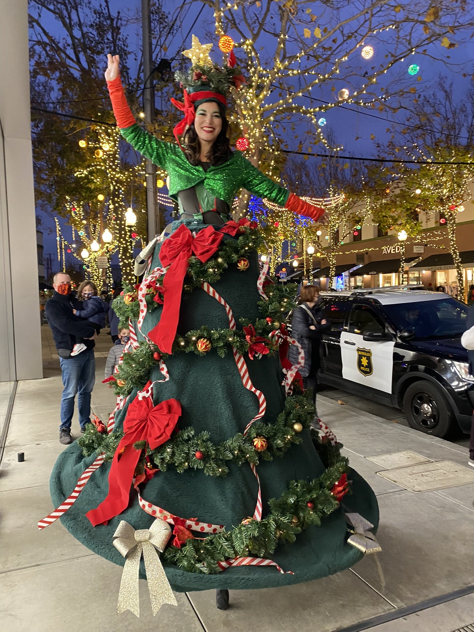 A woman dressed as an elf standing on a large decorated Christmas tree float, smiling, during a holiday parade at dusk, with Christmas lights on trees and people in the background.