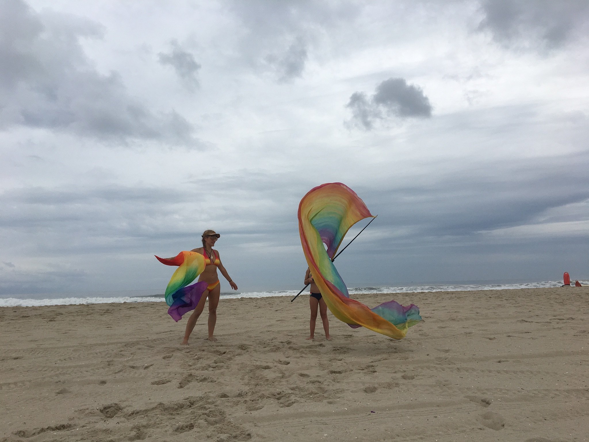 Two women on a beach, one holding a rainbow-colored flag and the other with a rainbow-colored scarf, under a cloudy sky, with ocean waves in the background.