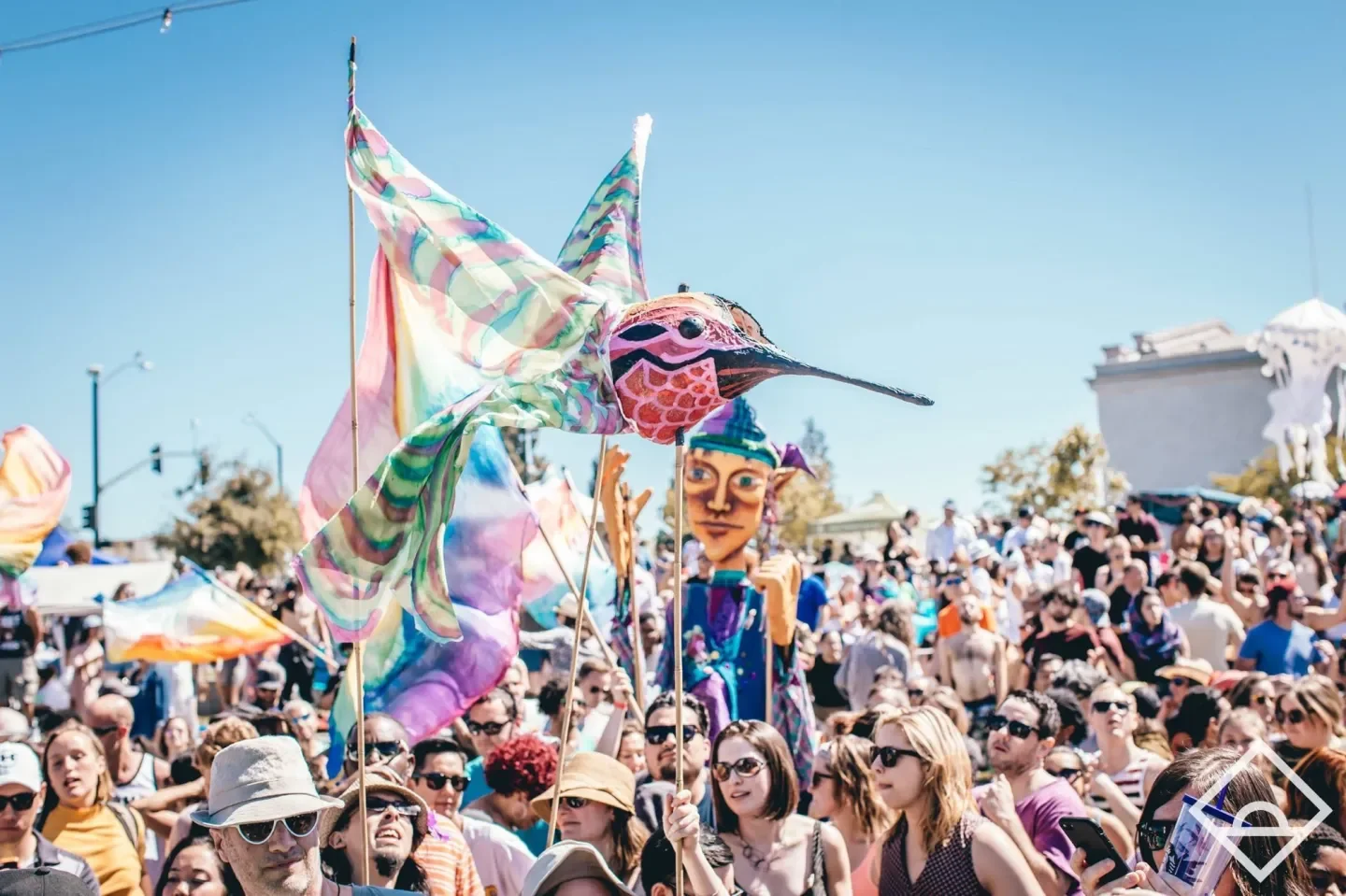 Crowd at a festival with people holding a colorful dragon kite and a large puppet head, sunny day with clear sky, some people wearing sunglasses and hats.