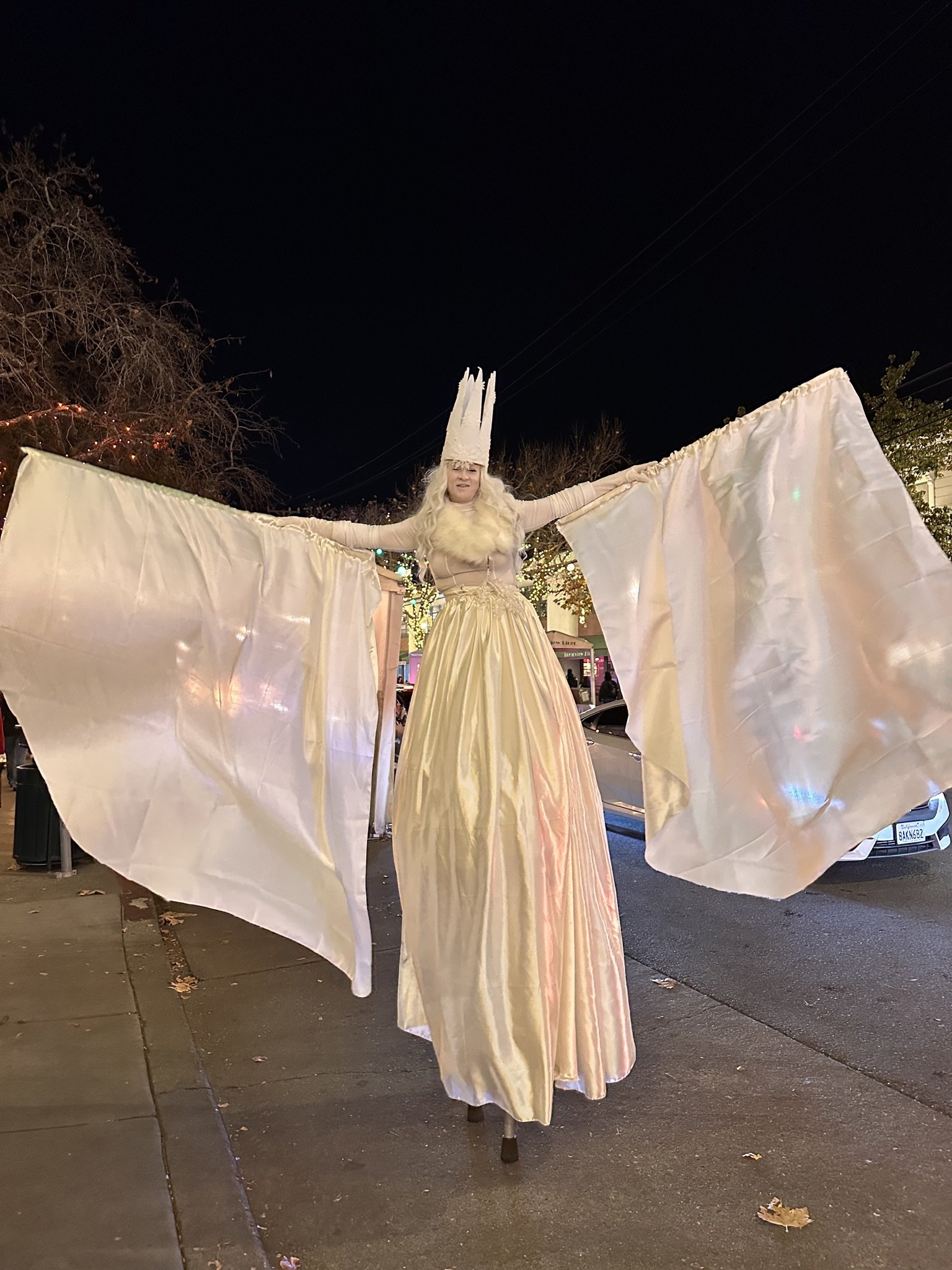 Person dressed as a Christmas angel or fairy with a tall white crown, long flowing dress, and large white wings, standing on stilts on a city sidewalk at night.