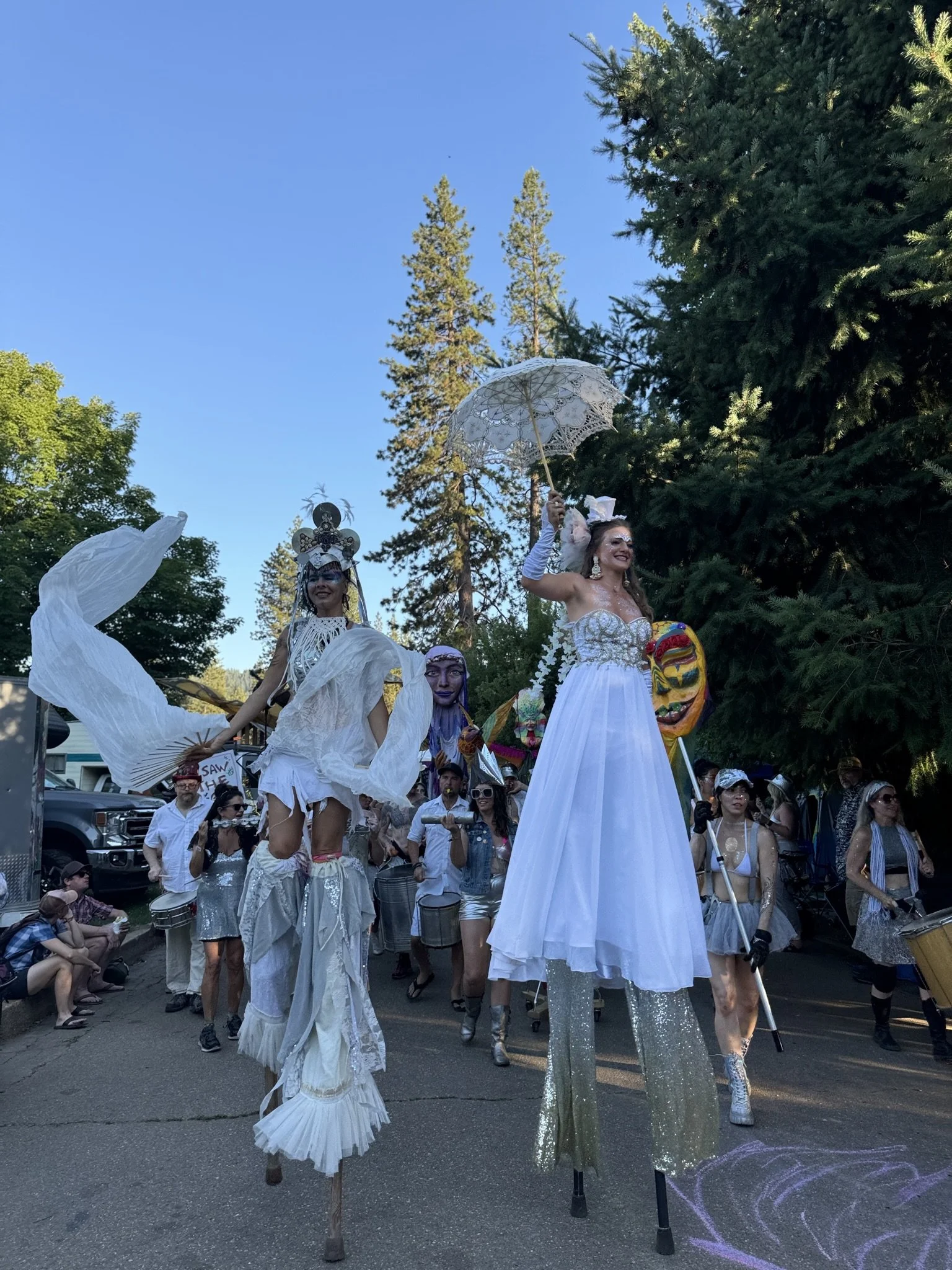 A parade featuring performers on stilts dressed in elaborate white costumes, some wearing masks and headpieces, with others holding umbrellas and musical instruments. Large trees and a crowd of spectators are visible in the background.