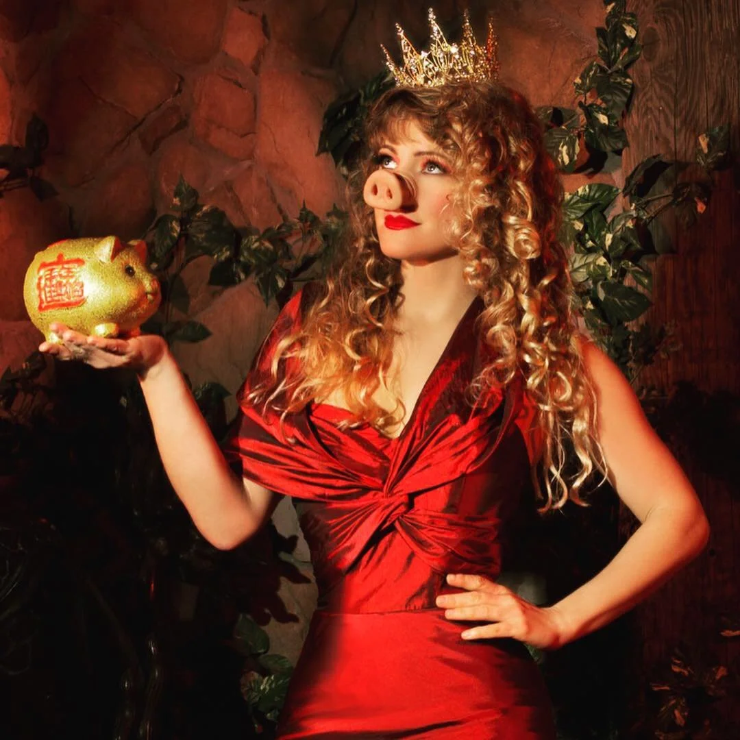 A woman with curly blonde hair dressed in a red dress wearing a crown, holding a piggy bank with Chinese characters on it, standing against a stone wall background with plants.