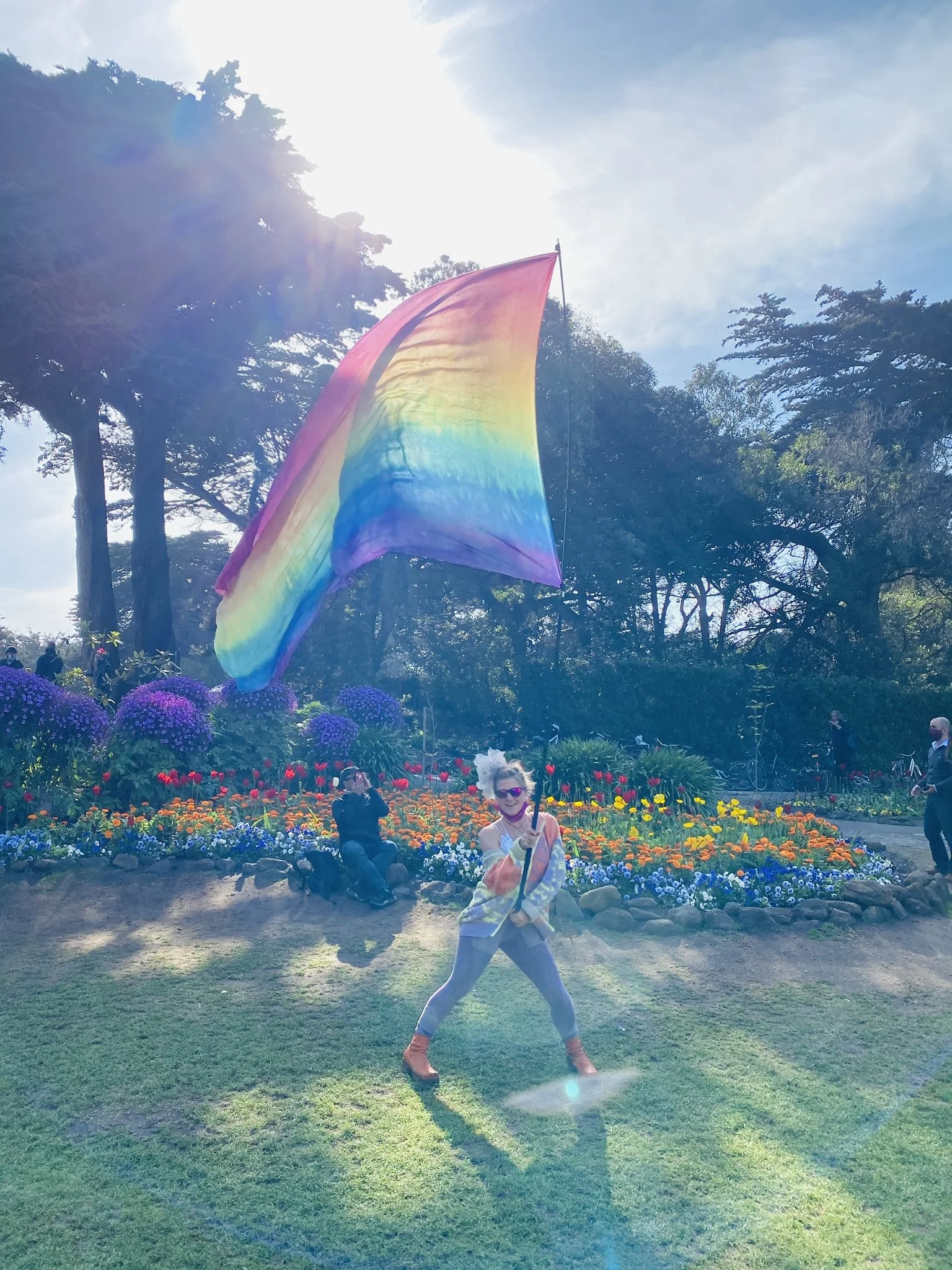 A woman wearing sunglasses, a colorful top, gray leggings, and orange boots is holding a pole with a large rainbow flag attached. She is standing on grass in front of a garden with purple, orange, and yellow flowers, with trees and several people in 