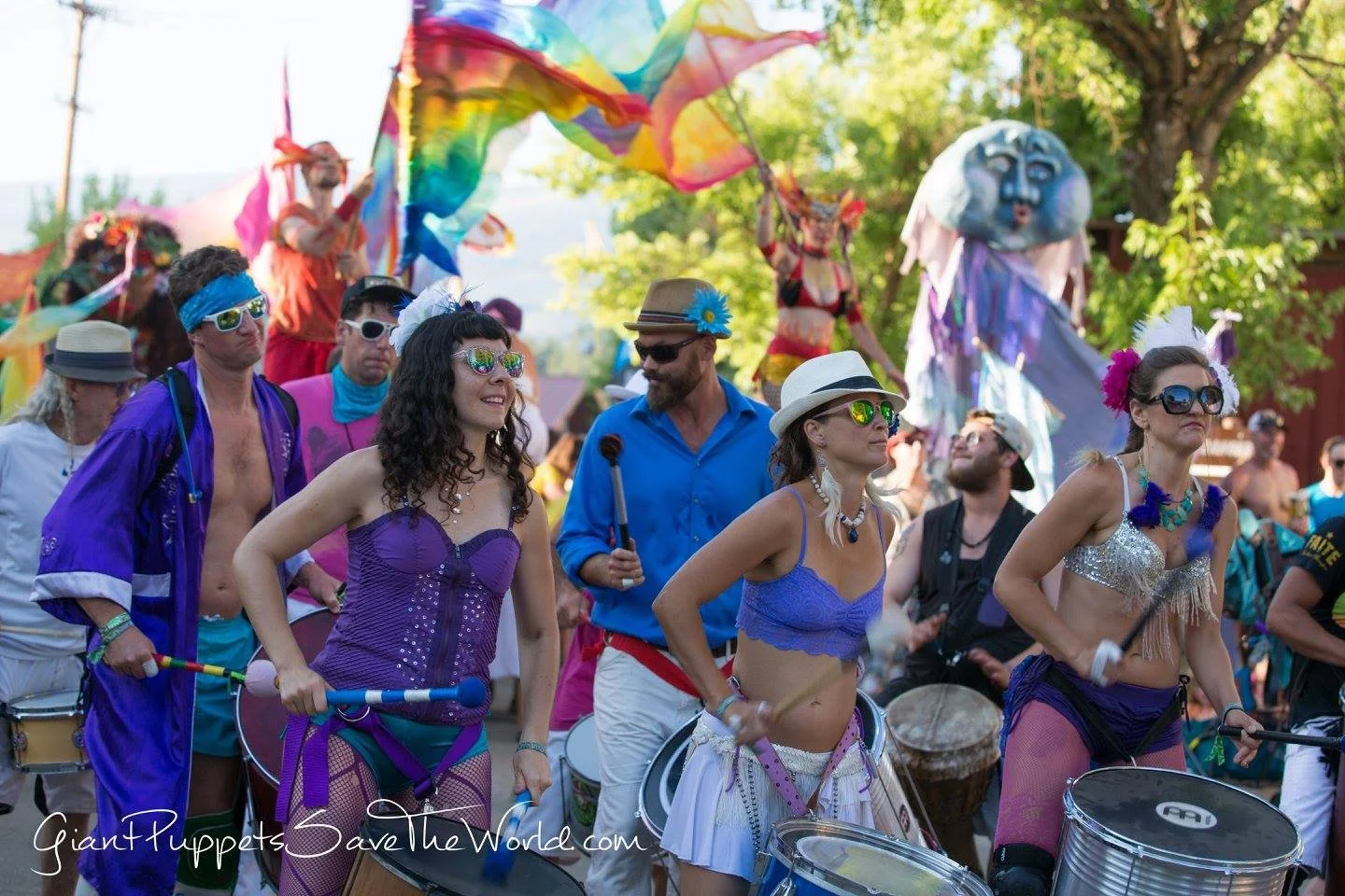 People at a parade wearing colorful costumes, some with drums, sunglasses, and accessories, with performers on stilts and large puppets in the background.