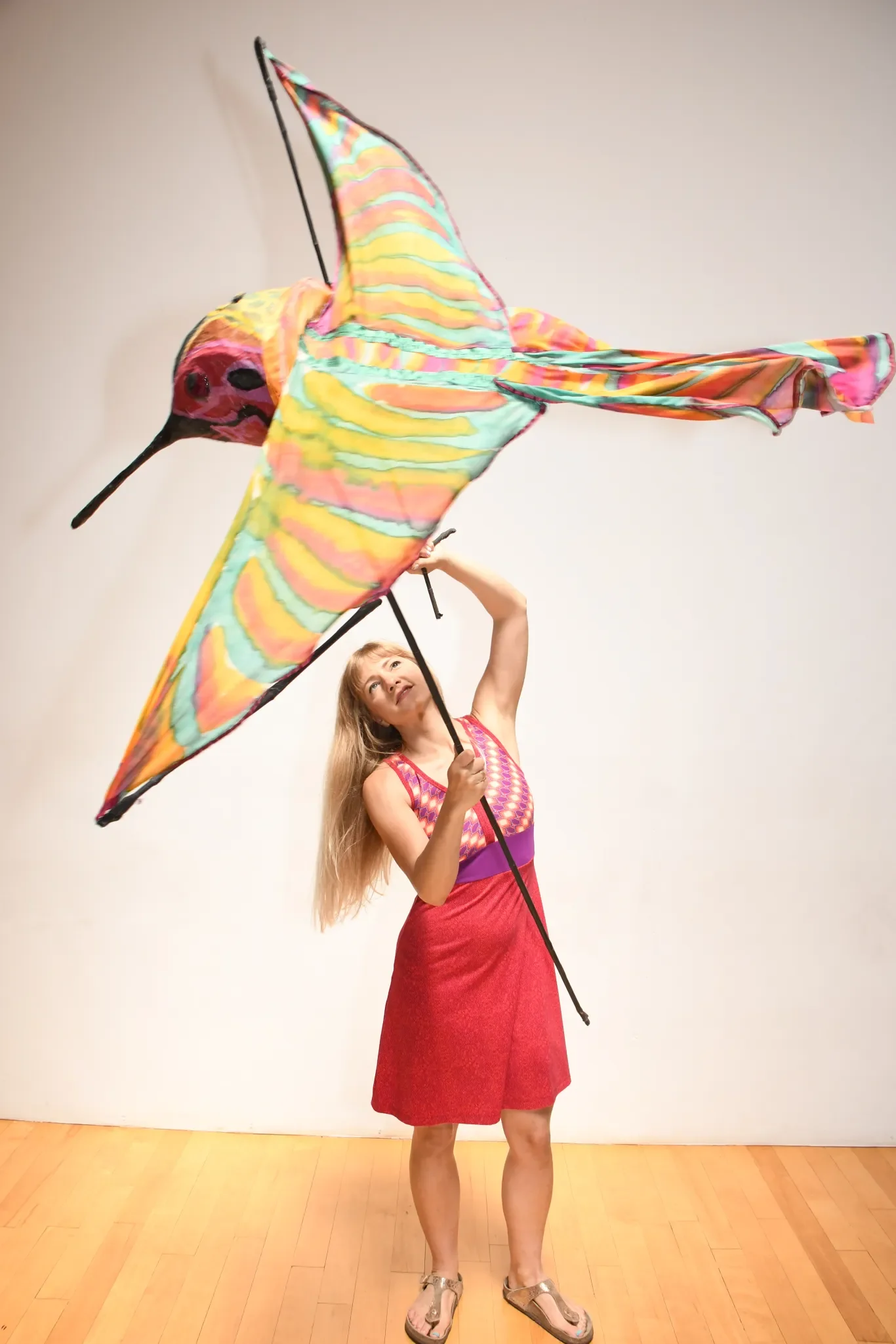 A woman holding a large colorful dragonfly-shaped kite indoors against a plain white wall and wooden floor.