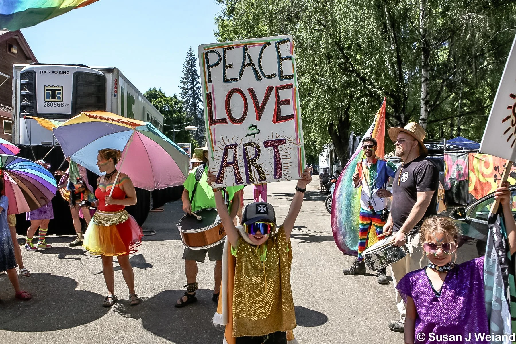 Children and adults participating in a colorful parade or festival, with one child holding a sign that says "PEACE LOVE ART," surrounded by vibrant umbrellas and costumes in a street lined with trees and buildings.