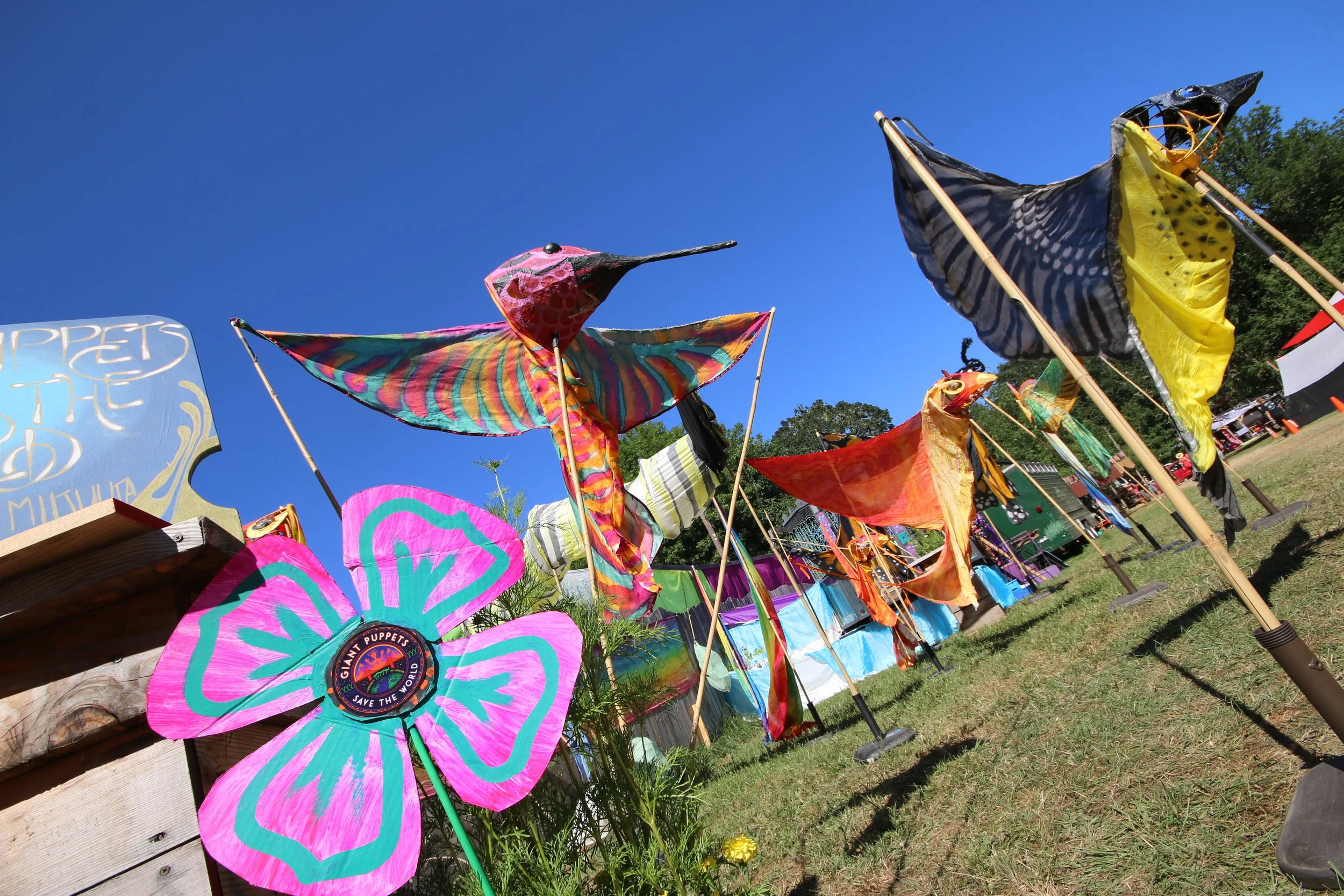Colorful fabric butterfly and bird puppets with vibrant wings and beaks are displayed on poles at an outdoor fair under a clear blue sky.