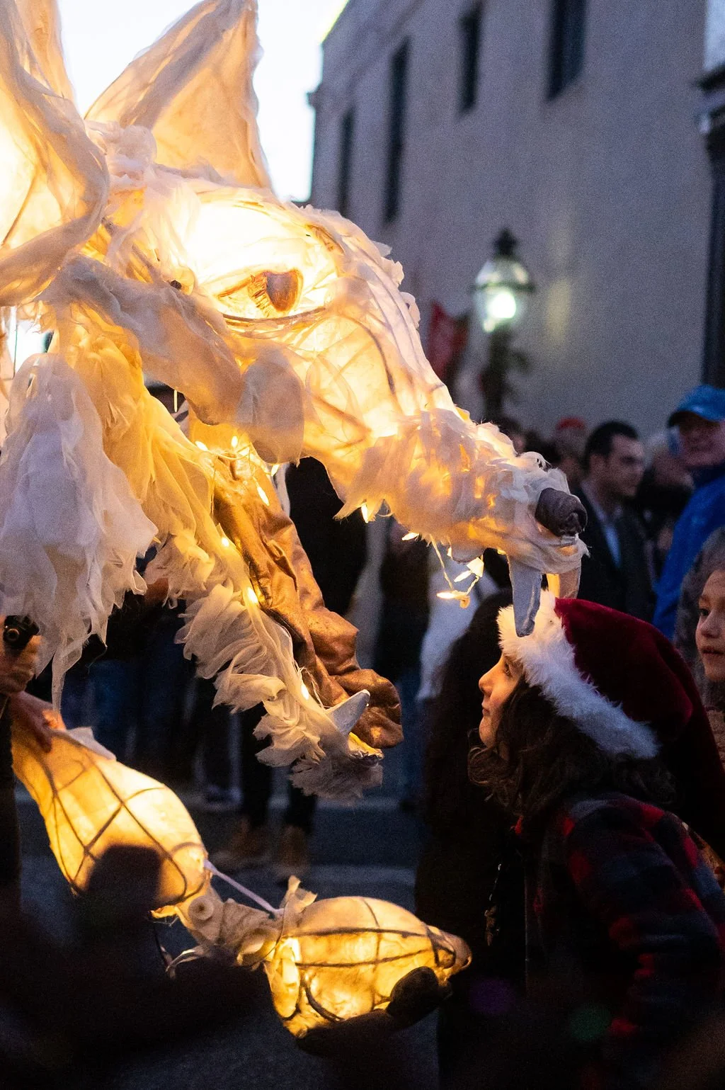 A girl wearing a Santa hat looks at a glowing dragon lantern at a holiday festival during the evening.