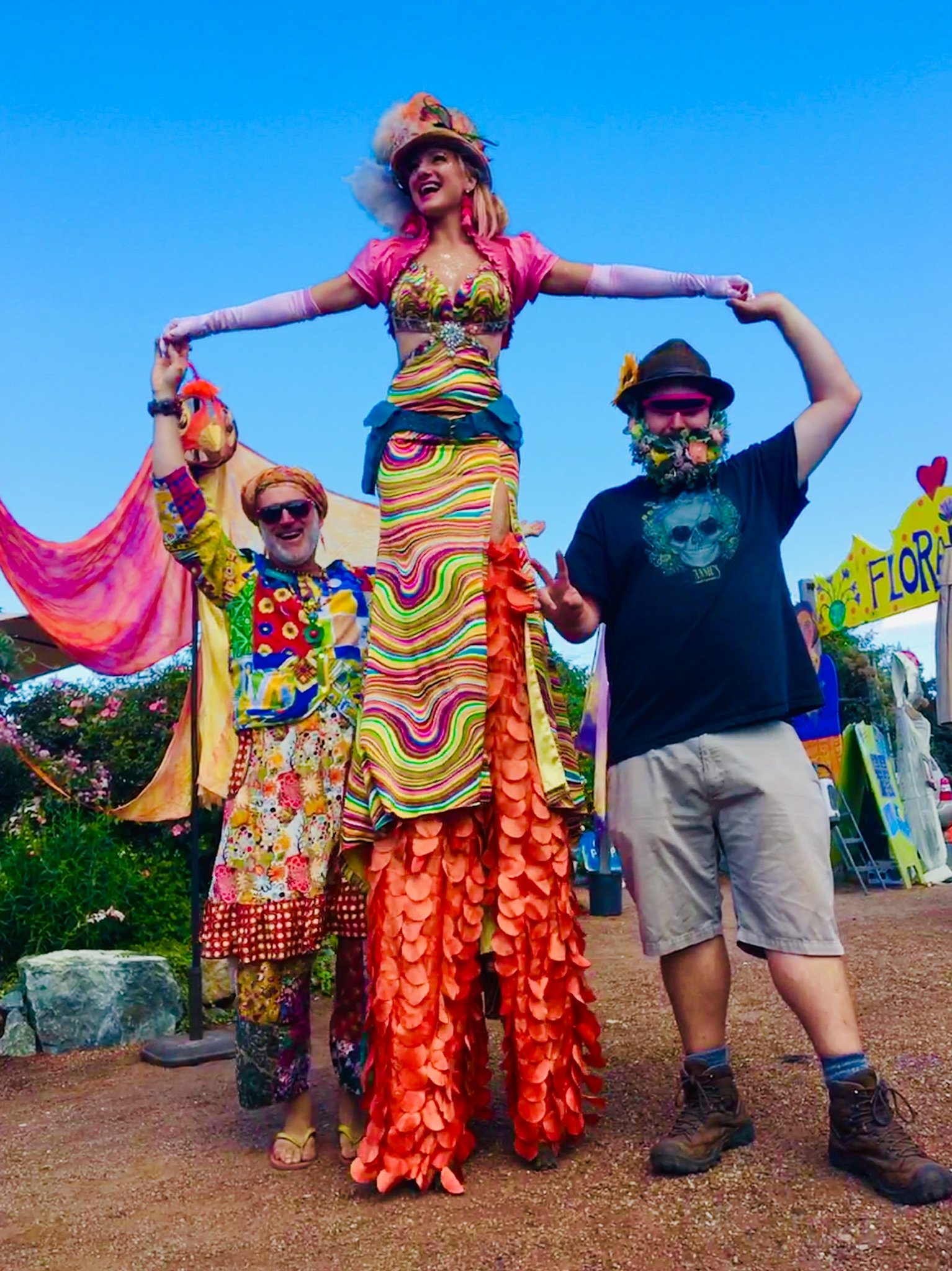 Three people wearing colorful clothing and costumes, one on stilts, in an outdoor festival setting with a clear blue sky.