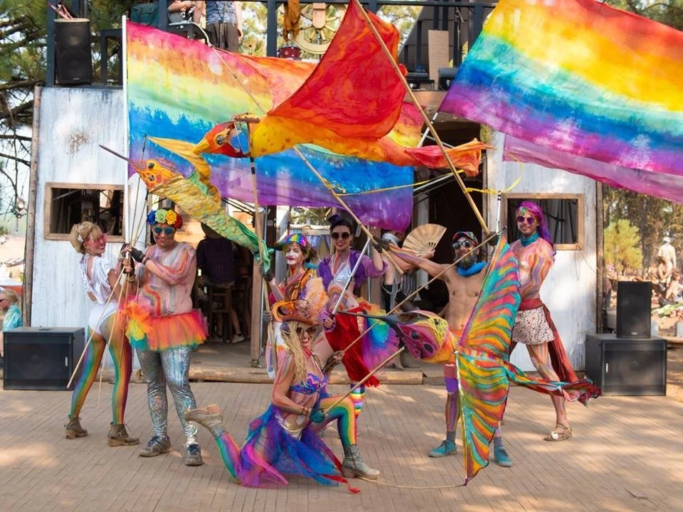 Group of performers dressed in colorful, rainbow-themed costumes holding large rainbow flags and fish-shaped puppets during a performance. The background shows a stage with a rustic wooden structure and some speakers.