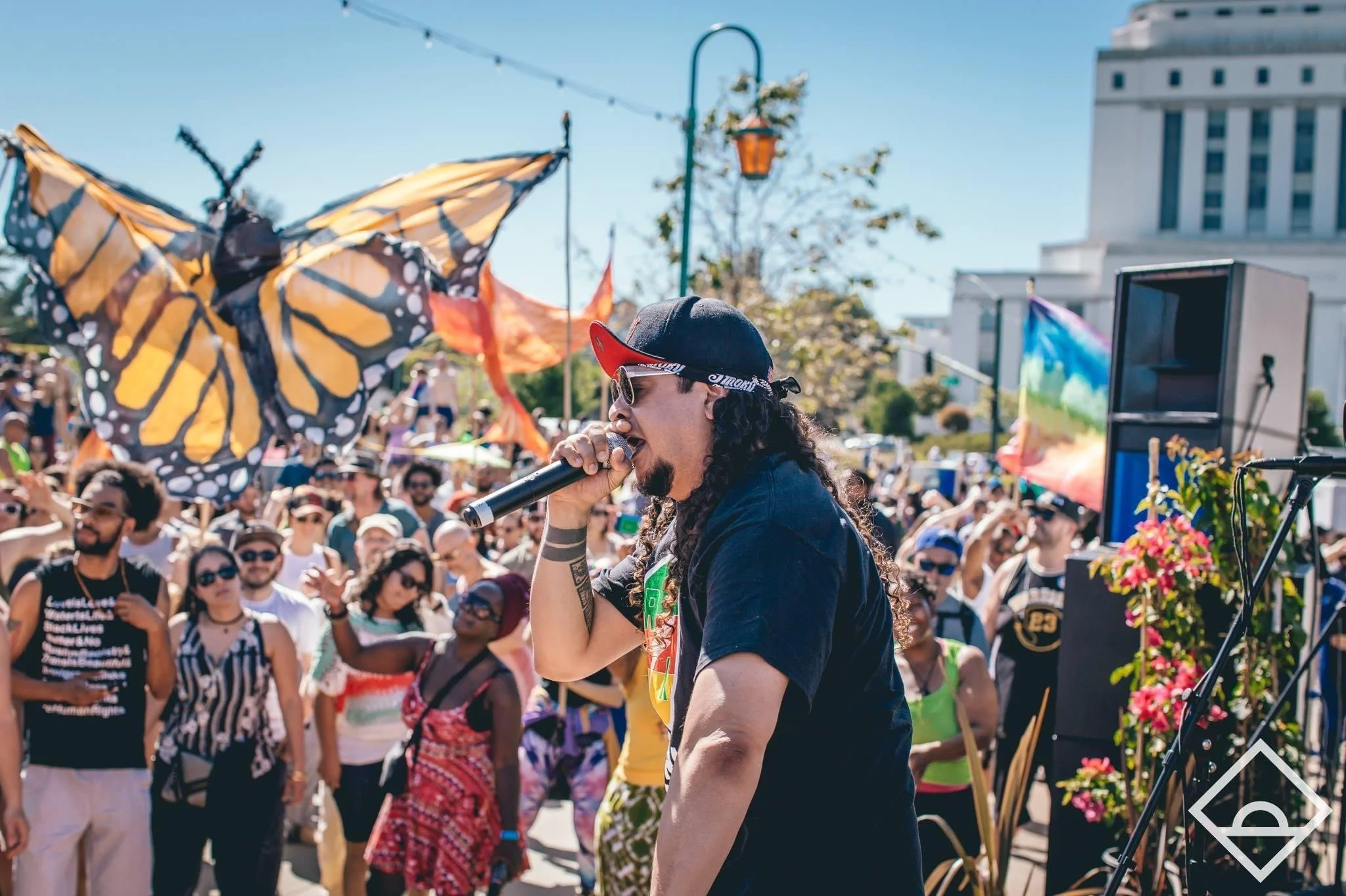 Performer singing into microphone at outdoor event with colorful butterfly decoration and crowd under bright sky.