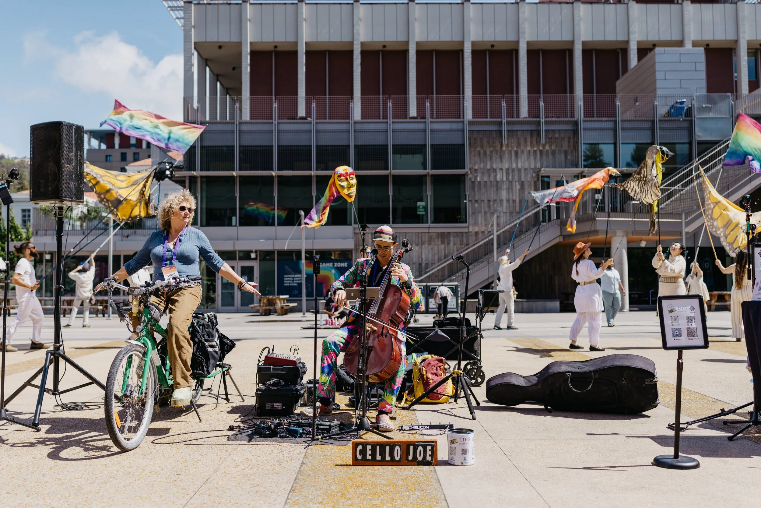 Street performers performing with flags and musical instruments, including a cello, in an outdoor urban plaza with a modern building in the background.