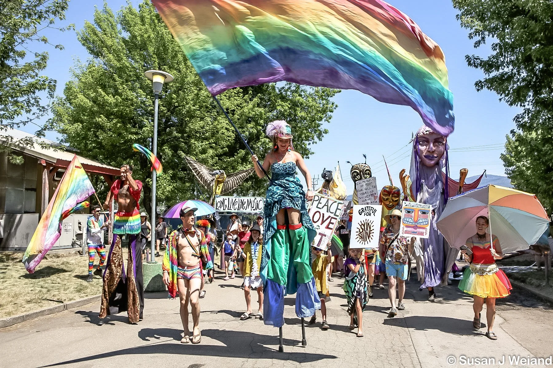 A lively LGBTQ pride parade with people dressed in colorful outfits, holding rainbow flags, posters, and umbrellas. There are large artistic figures and signs advocating for peace, love, and equality. The parade takes place outdoors on a sunny day wi