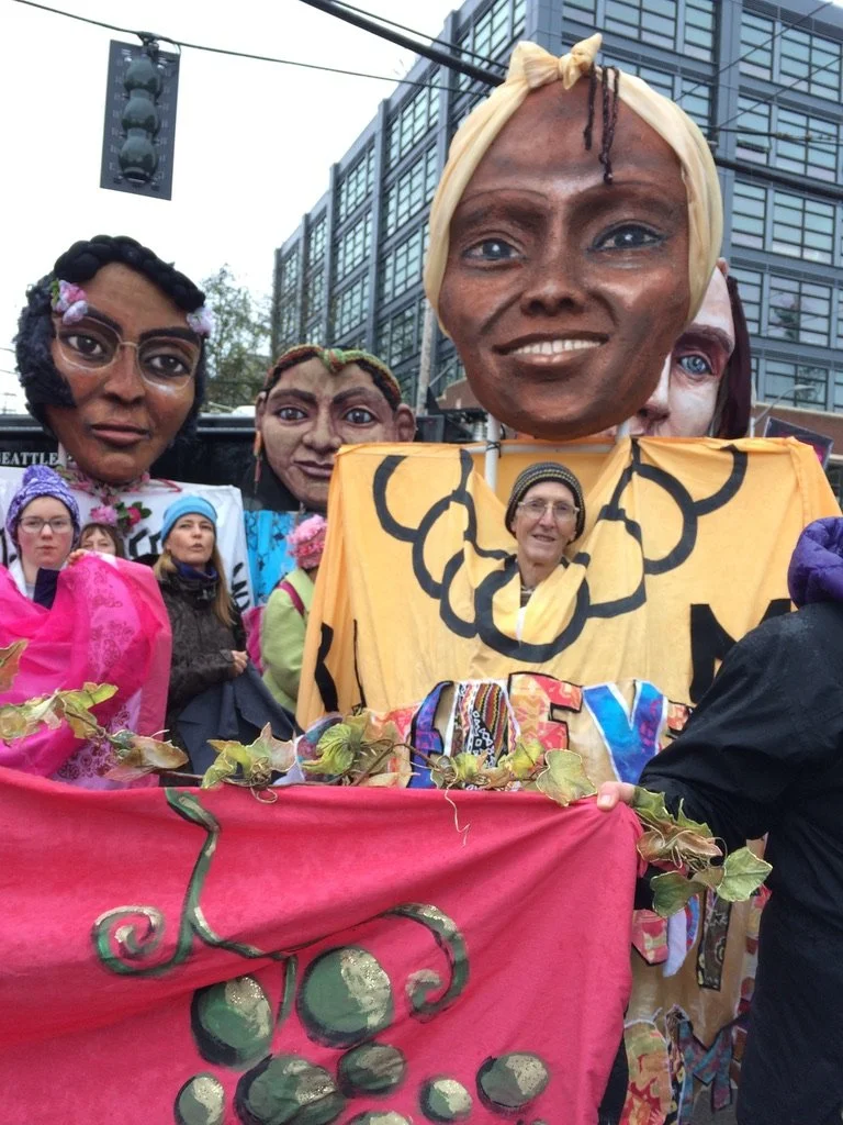 Group of people holding large colorful puppets of diverse women's faces, some adorned with flowers and headscarves, in an outdoor urban setting.