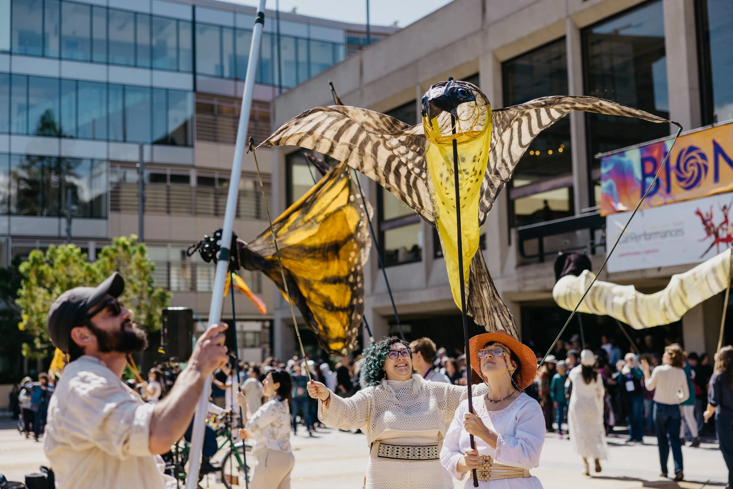 People participating in an outdoor event or parade holding large, colorful butterfly-shaped kites in the air, with a crowd and modern buildings in the background.